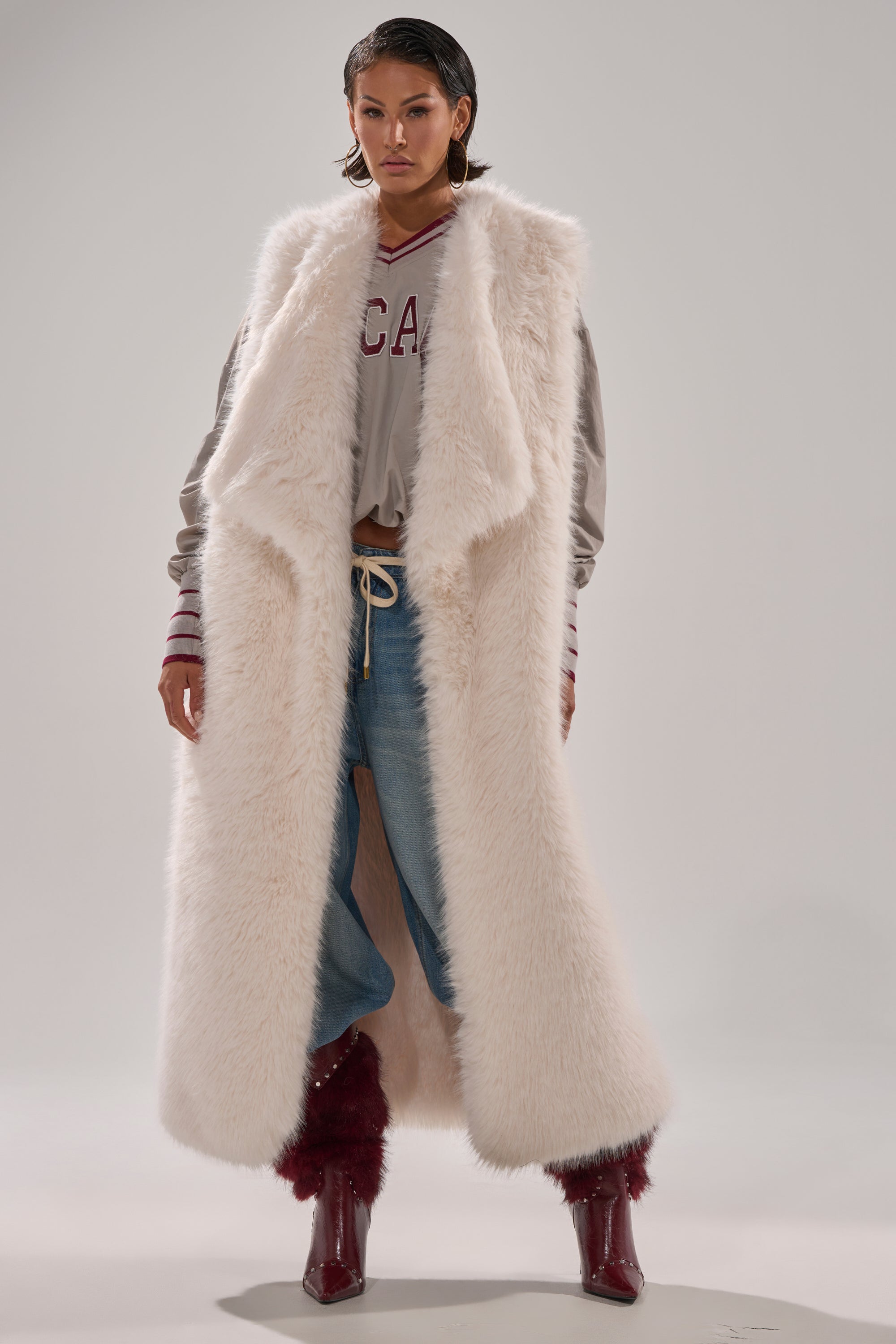 Wearing the DOWN TIME FAUX FUR VEST IN IVORY over a gray sweatshirt and loose light blue jeans, paired with maroon furry boots and wet-look hair, a person stands against a plain light background. 0
