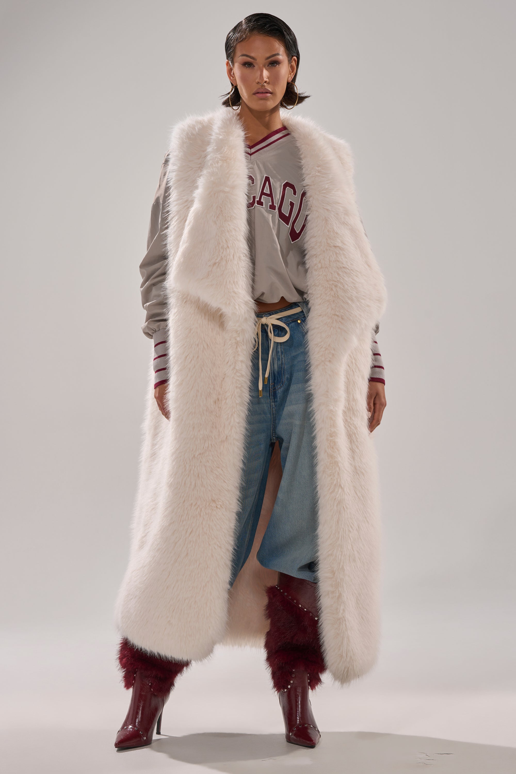 A woman stands confidently in the DOWN TIME FAUX FUR VEST IN IVORY over a gray "CHICAGO" sweater, paired with a front-slit denim skirt and red boots against a plain background. 1