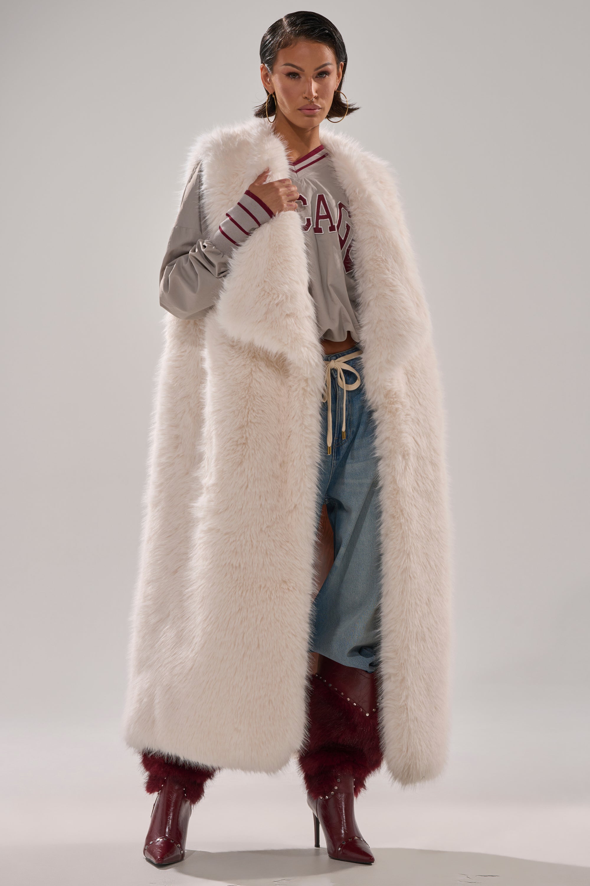 A woman stands confidently against a plain background wearing the DOWN TIME FAUX FUR VEST IN IVORY over a gray sweater with red text, distressed blue jeans, and burgundy boots. 2