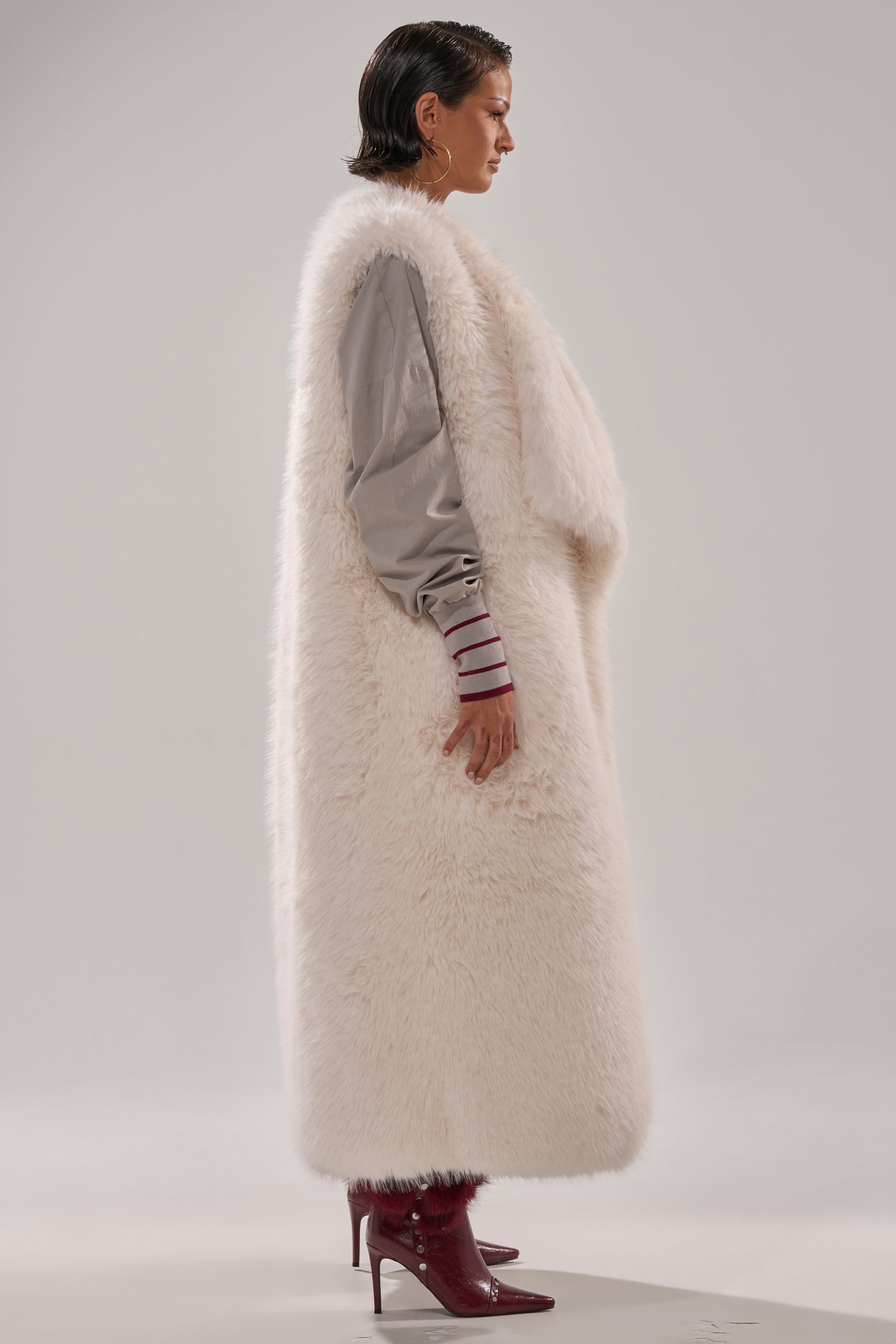 A person stands in profile wearing the DOWN TIME FAUX FUR VEST IN IVORY over a gray top with striped cuffs and burgundy high-heeled boots, set against a plain light background. 3