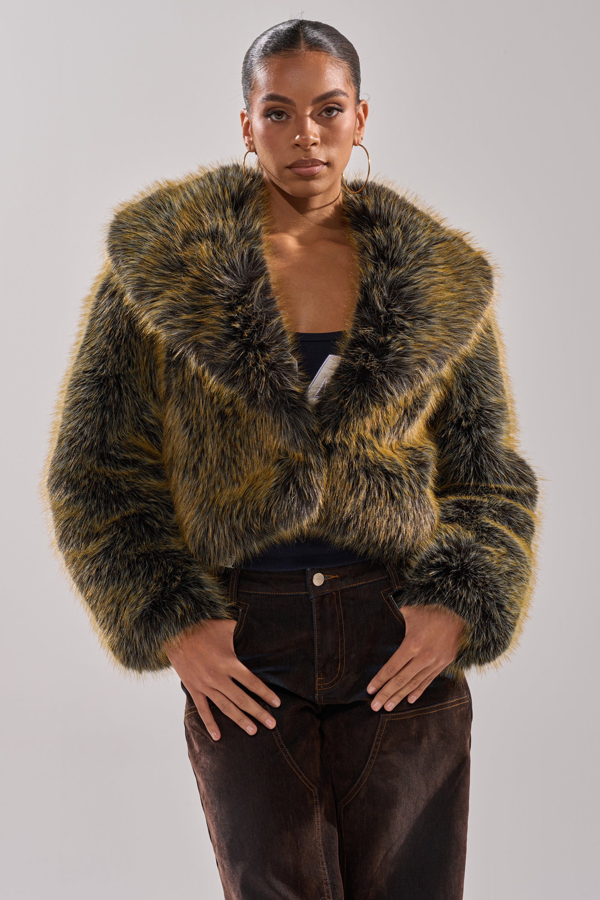 A woman stands facing forward in the LIMONCELLO FAUX FUR COAT, featuring brown and yellow tones with an oversized shawl collar. Her hair is pulled back, hoop earrings shine, and she exudes confidence against a plain background.