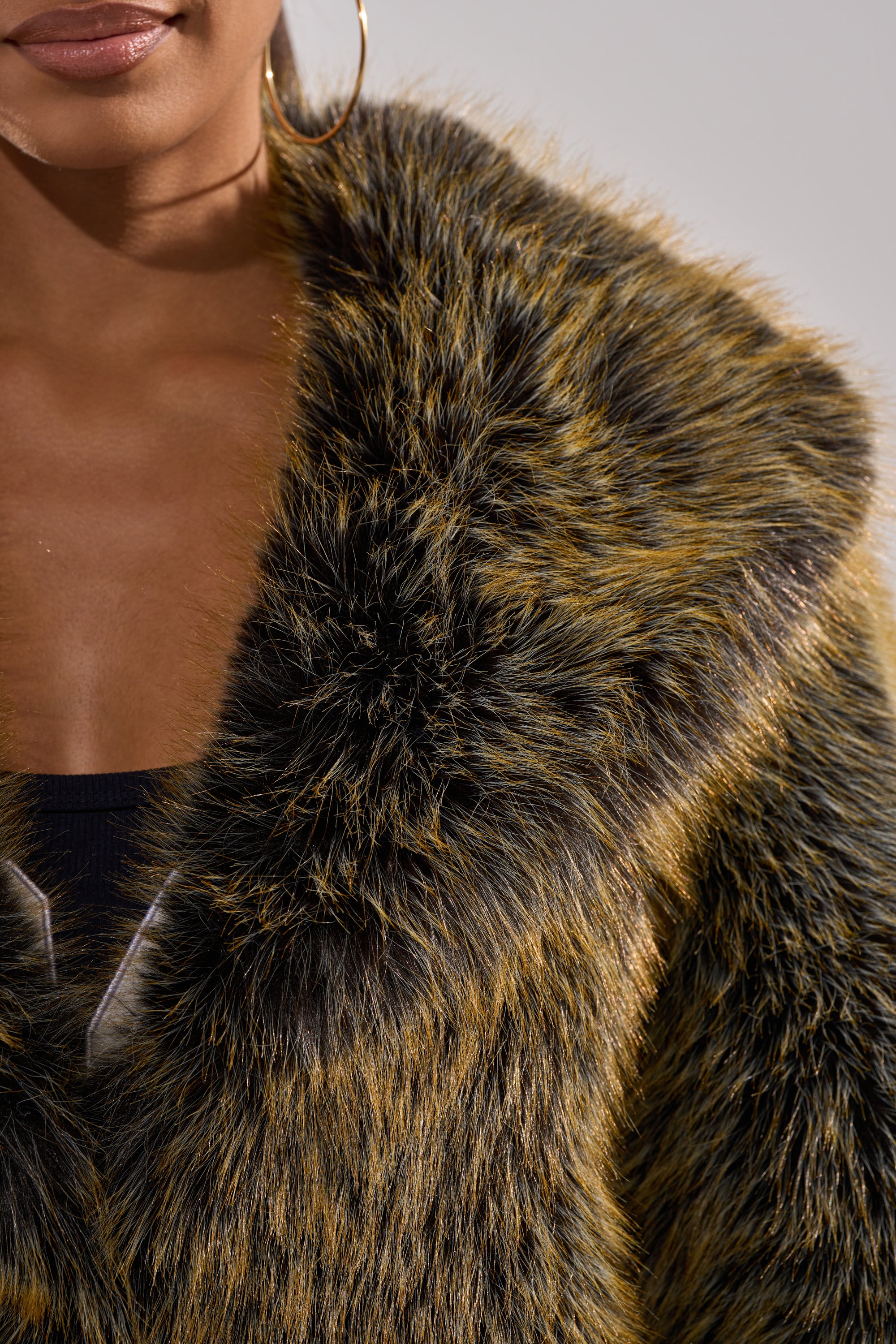 A close-up of a woman wearing the LIMONCELLO FAUX FUR COAT with black and golden brown streaks, an oversized shawl collar, paired with a black top and large hoop earring.