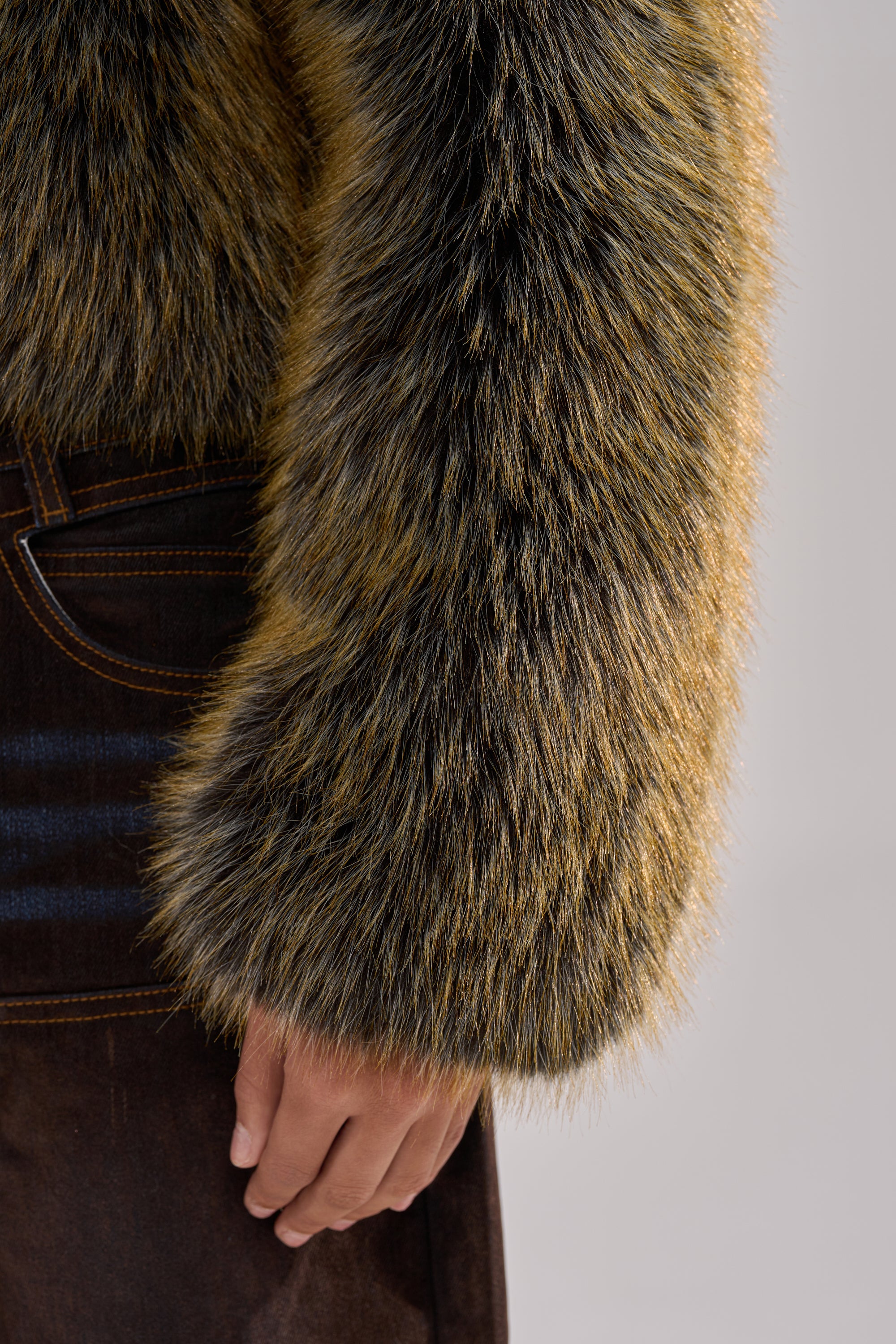 A close-up shows a person wearing the LIMONCELLO FAUX FUR COAT with an oversized shawl collar and boxy, waist-length fit, styled with dark brown pants. Their sleeve and hand rest by their side against a blurred neutral background.