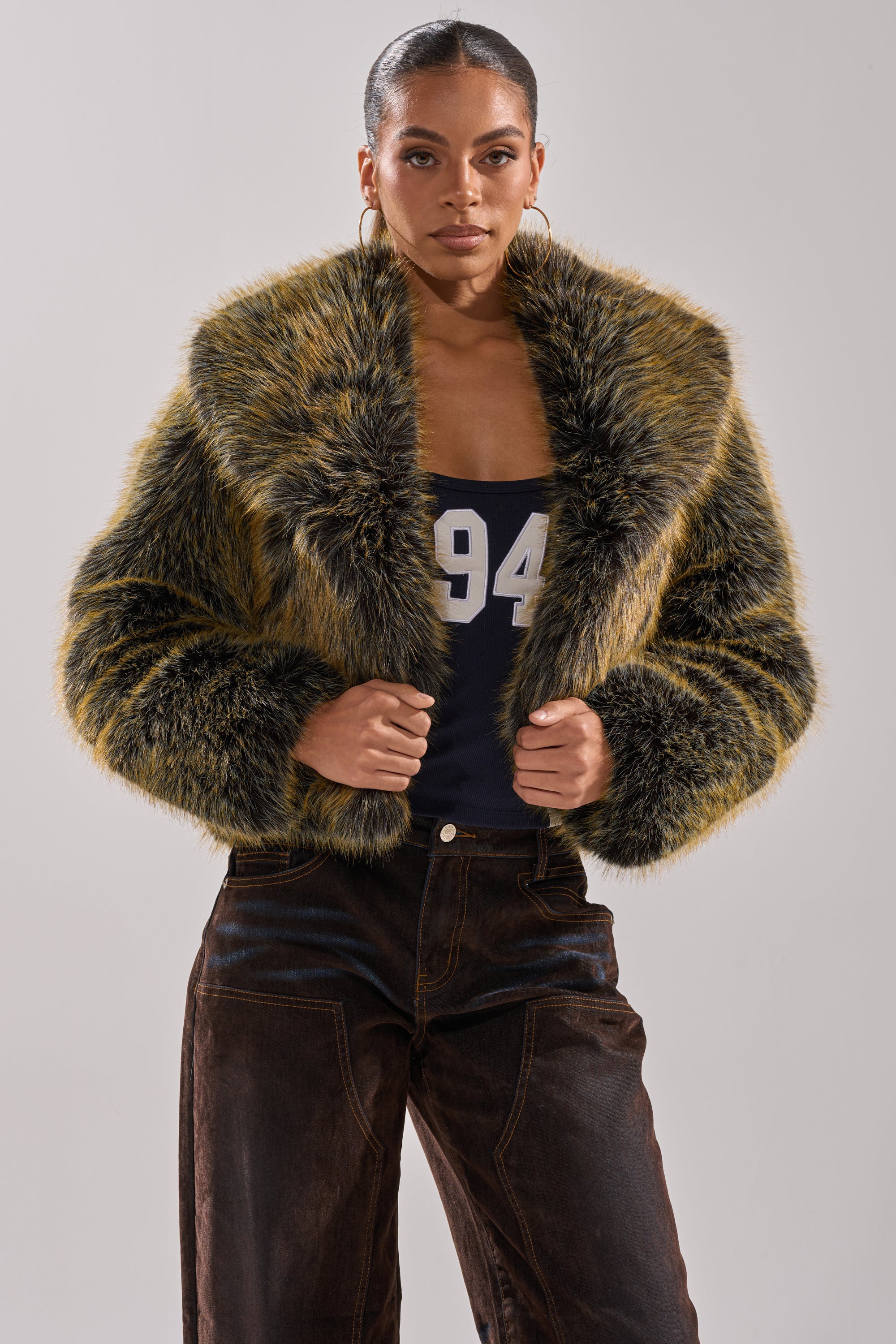 A woman stands confidently in the LIMONCELLO FAUX FUR COAT, a yellow and black design with an oversized shawl collar, layered over a navy top with "94" and dark brown pants against a plain light background.