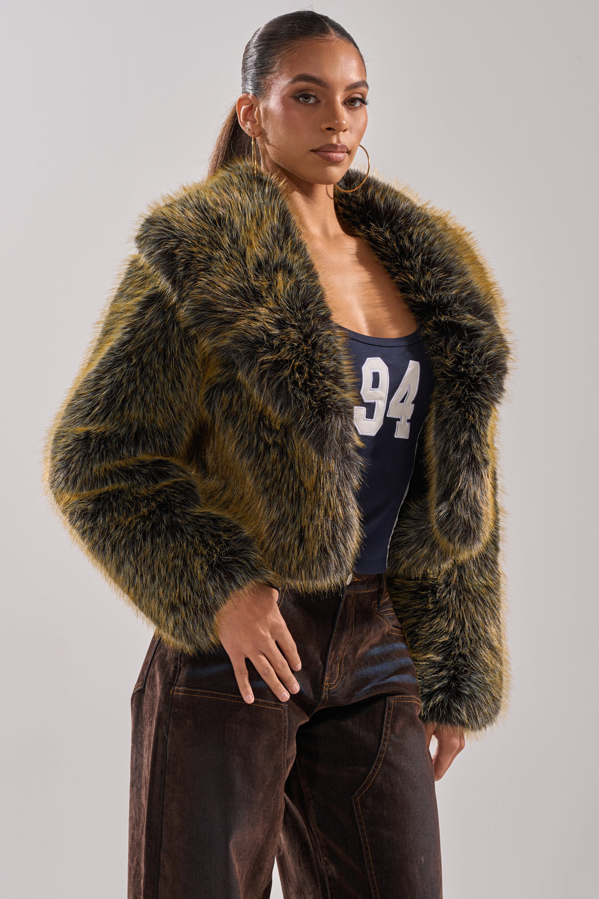A woman poses confidently in the LIMONCELLO FAUX FUR COAT, featuring a furry, oversized shawl collar in brown and black hues, styled over a navy top with "94" and high-waisted dark brown pants against a plain backdrop.