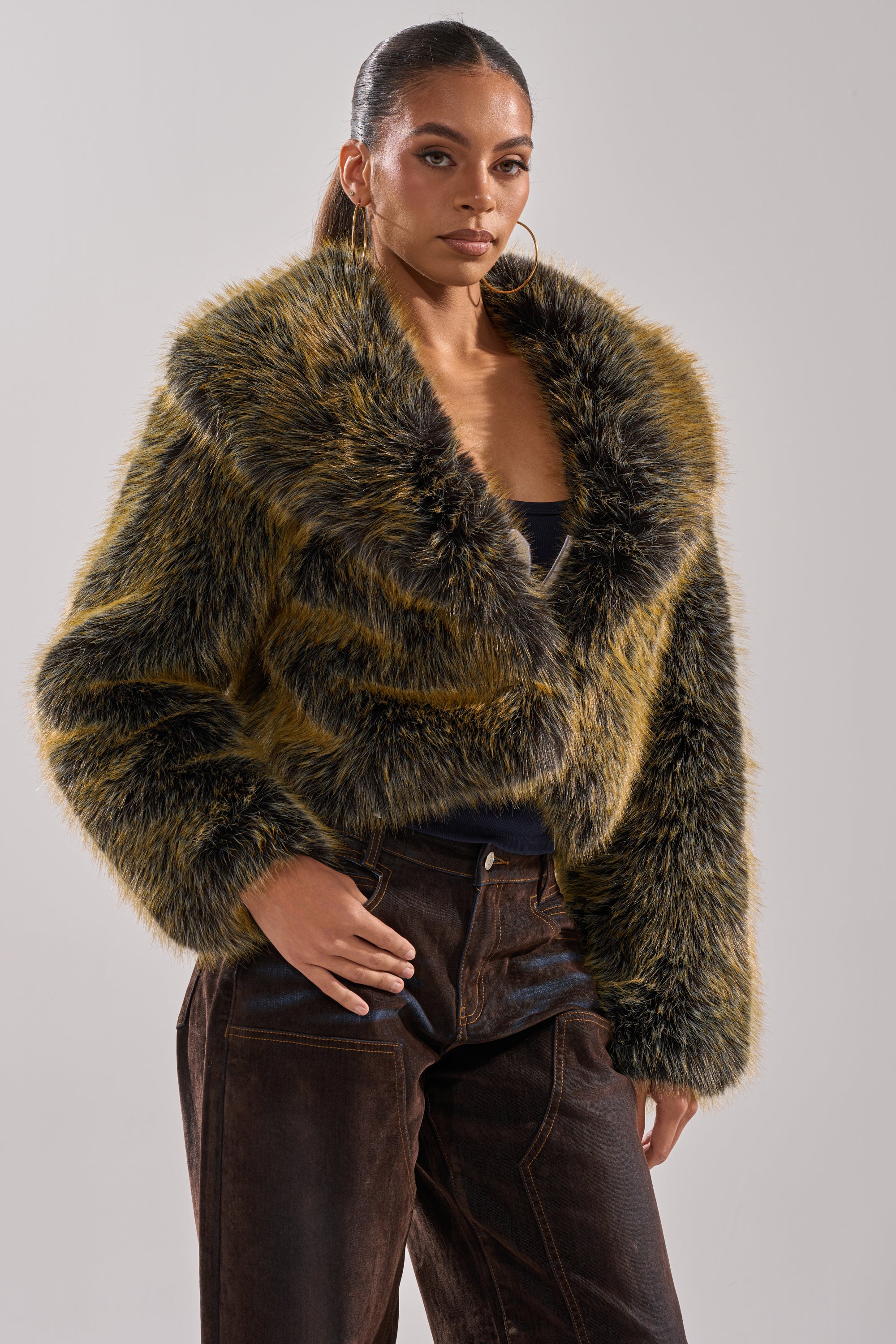 A woman models the LIMONCELLO FAUX FUR COAT—an oversized jacket with a wide collar—over a black top and brown pants, standing against a plain, light background.