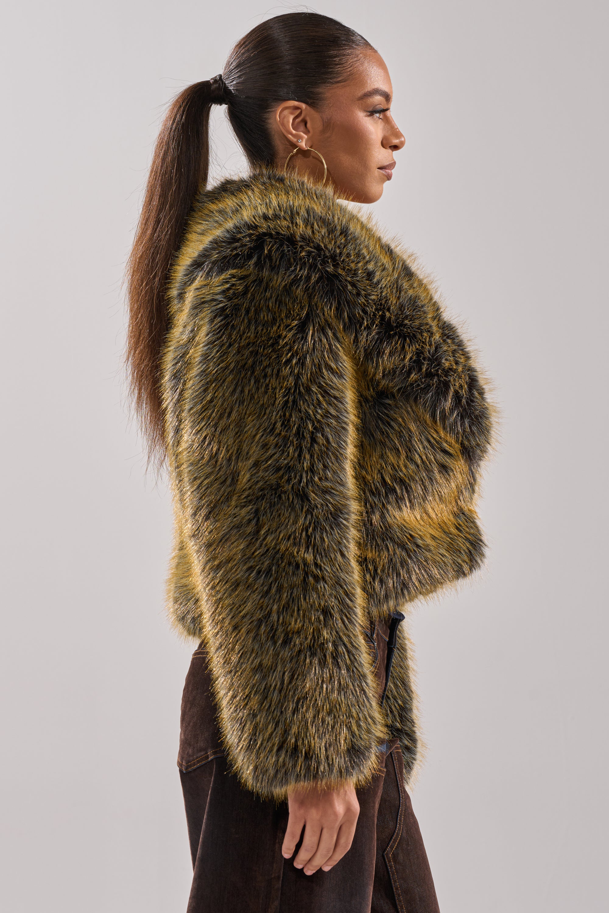 A woman with long straight hair in a ponytail stands sideways against a plain light background, wearing dark pants and the oversized yellow-and-black LIMONCELLO FAUX FUR COAT.