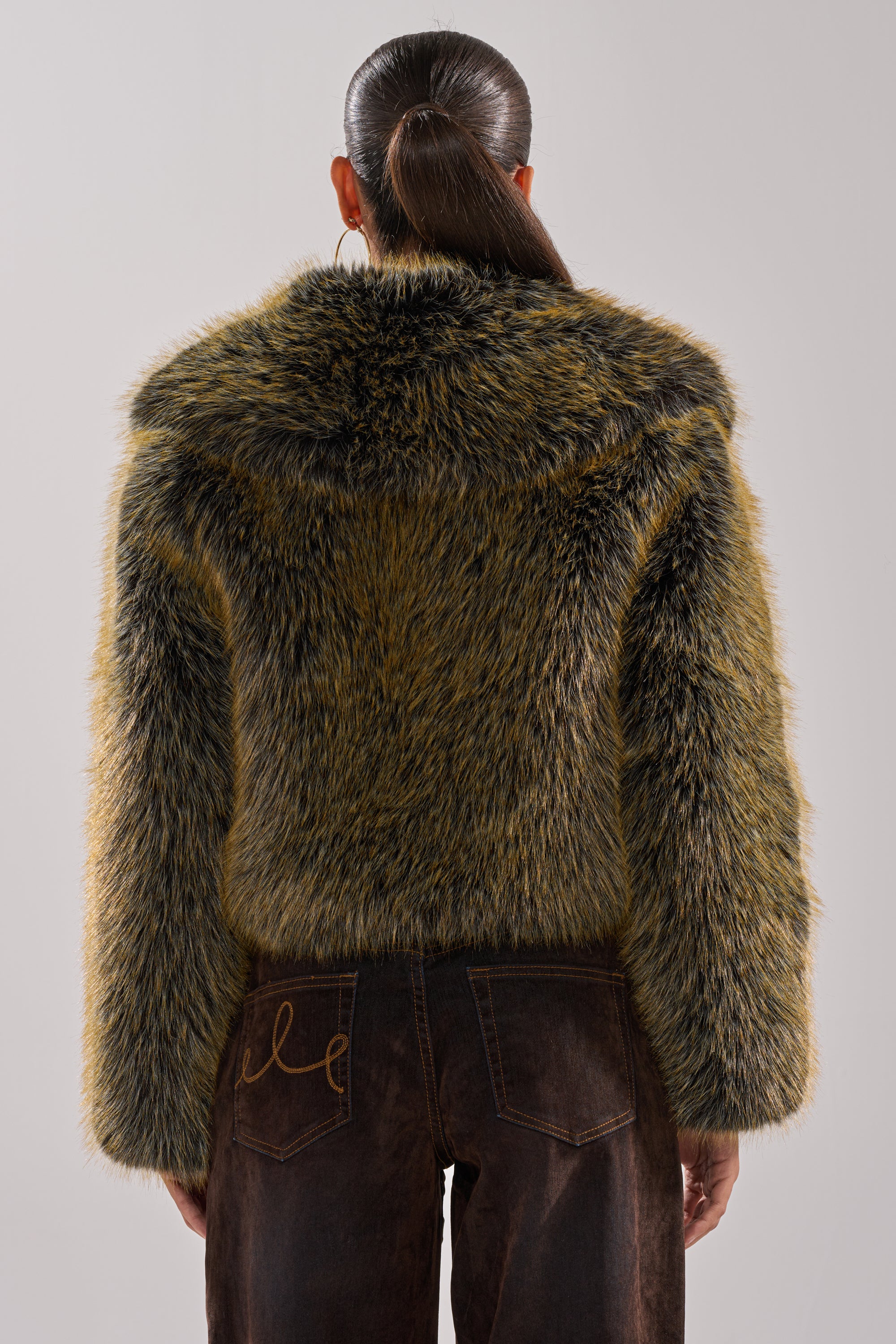 A person with long dark hair in a low ponytail is seen from behind wearing the oversized LIMONCELLO FAUX FUR COAT in brown and olive, paired with dark brown jeans, set against a plain light background.