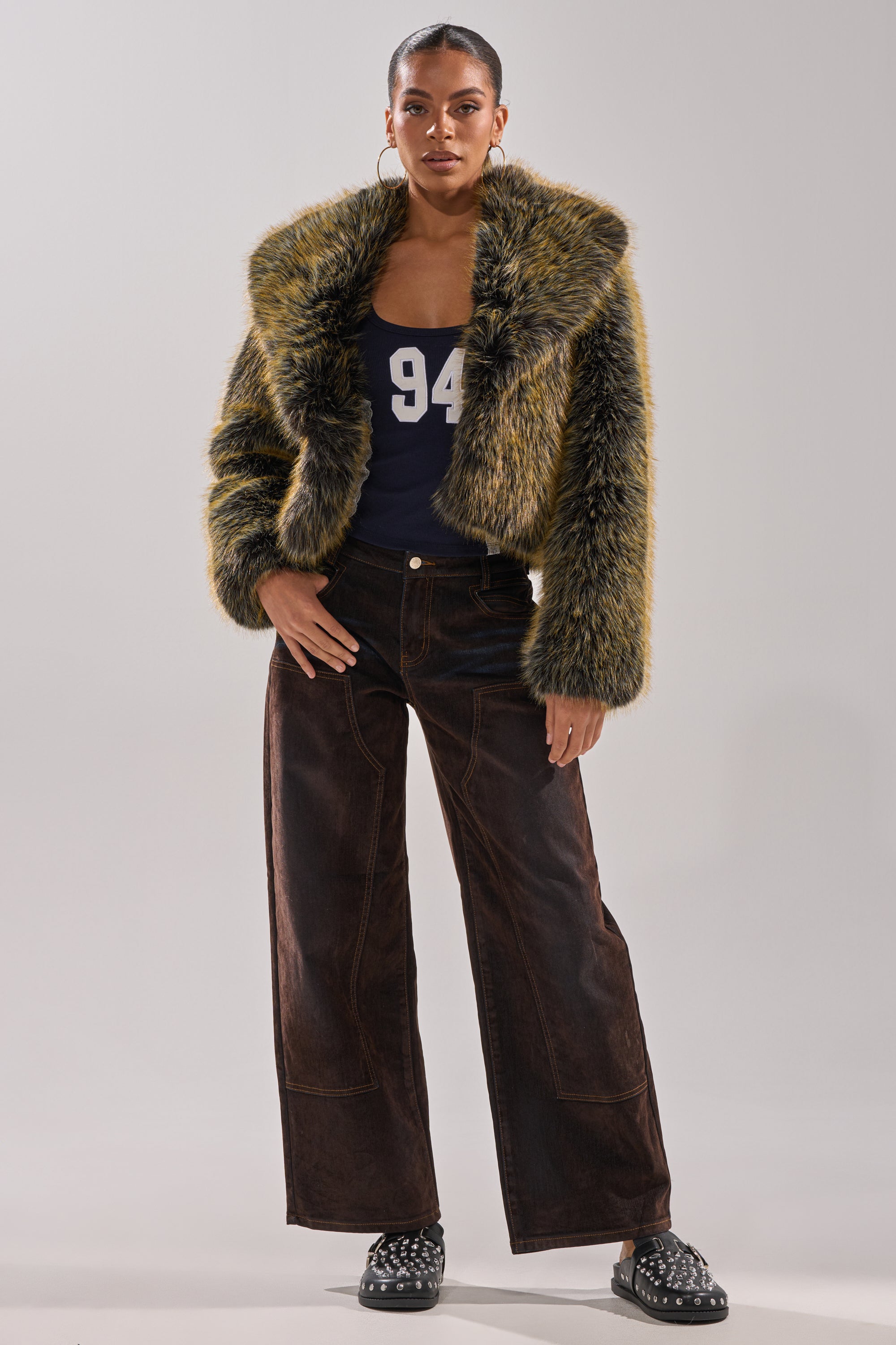 A woman faces forward in the LIMONCELLO FAUX FUR COAT—yellow and black with an oversized shawl collar—paired with a navy "94" tank, brown wide-leg pants, black studded shoes, hoop earrings, and her hair pulled back.