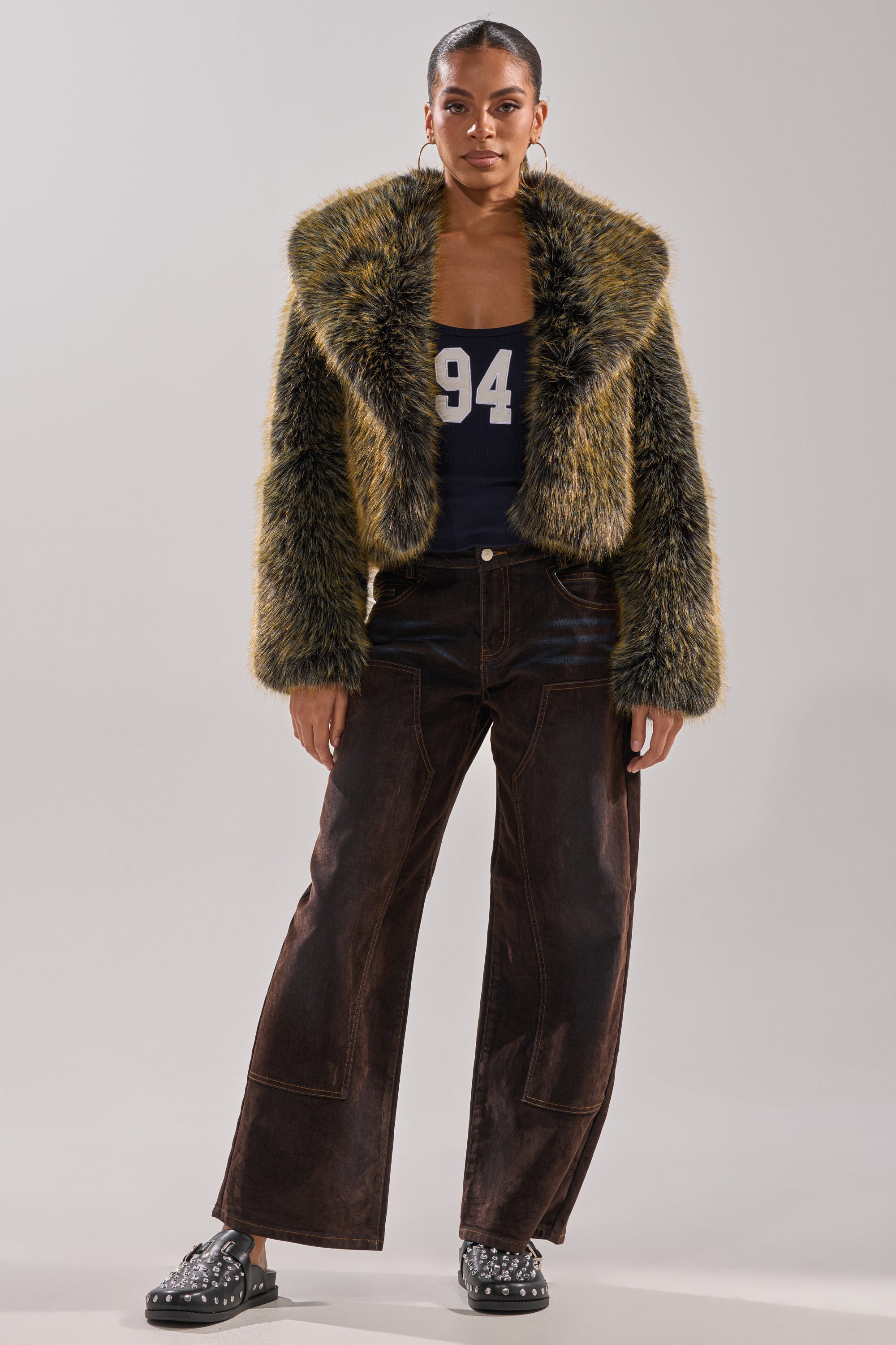 A woman stands wearing the LIMONCELLO FAUX FUR COAT with an oversized shawl collar, a navy "94" tank top, wide-leg brown pants, and black studded slip-on shoes. Her hair is sleeked back and her makeup is neutral.