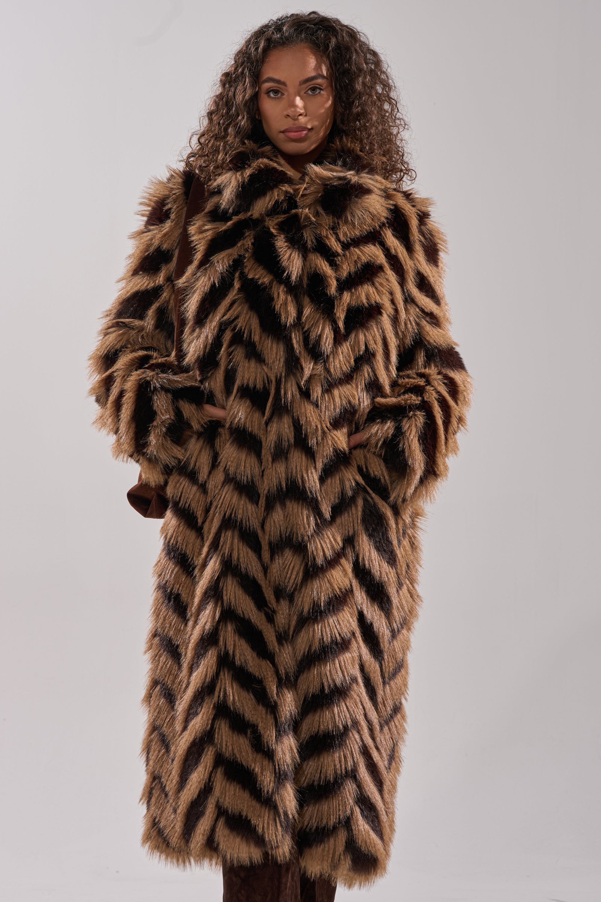 A woman with curly hair models the AUTUMN FAUX FUR COAT, a longline coat with a bold brown and black chevron pattern, standing against a plain light background.