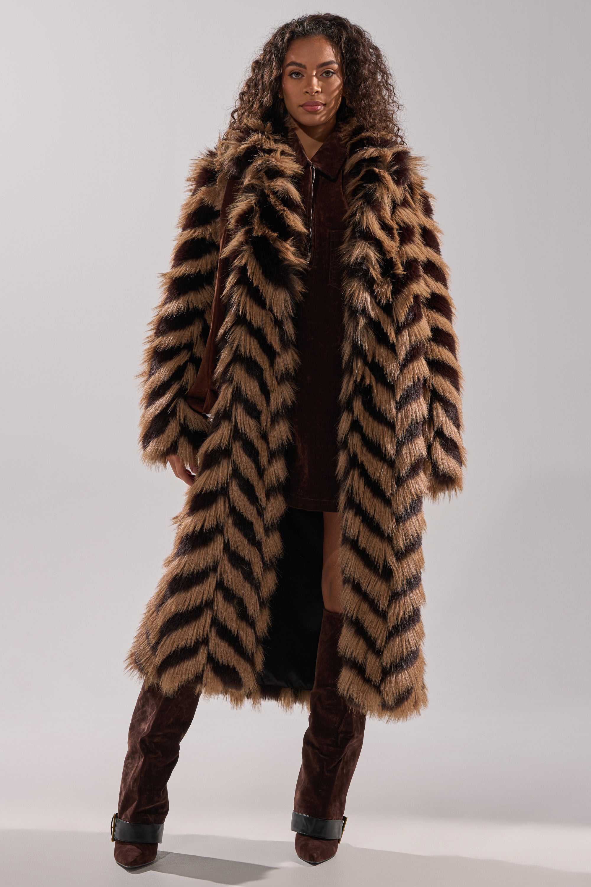 A woman models the AUTUMN FAUX FUR COAT, featuring a longline chevron print, layered over a dark brown outfit with tall brown boots against a neutral studio background.
