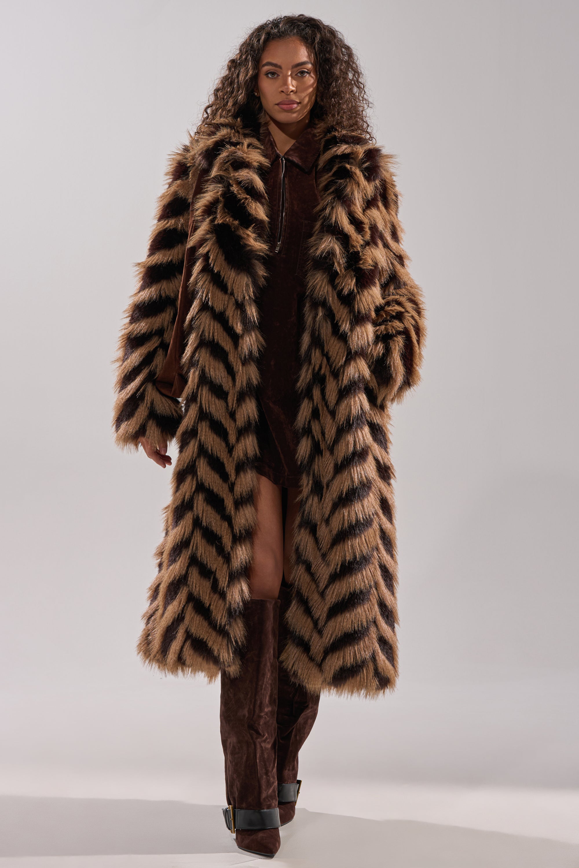A woman models the AUTUMN FAUX FUR COAT, featuring brown and black stripes, layered over a brown dress and knee-high boots, posing against a plain light background.