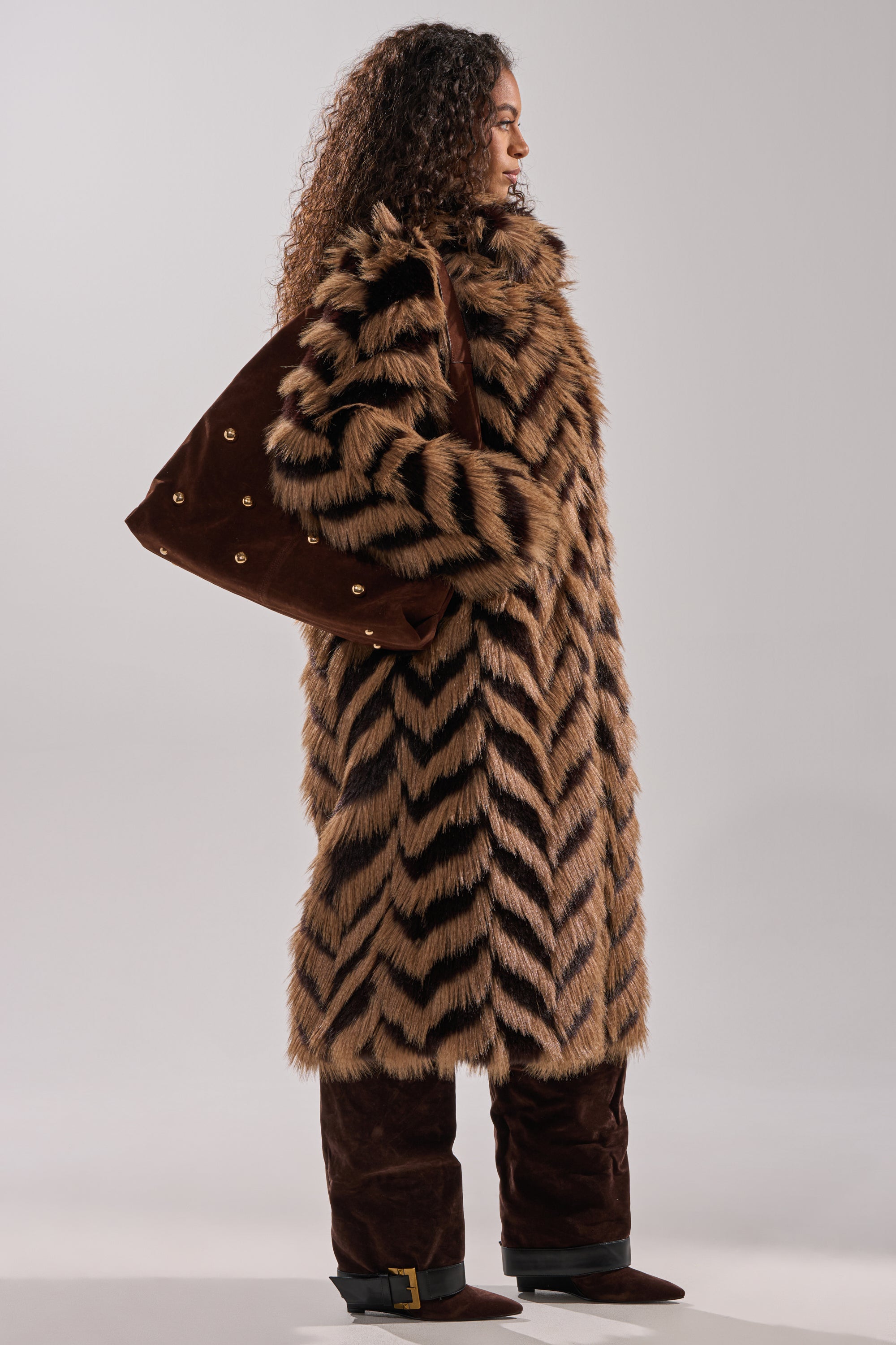 A person with curly hair stands in profile, wearing the AUTUMN FAUX FUR COAT featuring a brown and black chevron pattern, paired with brown pants, boots, and carrying a large brown bag with gold details.