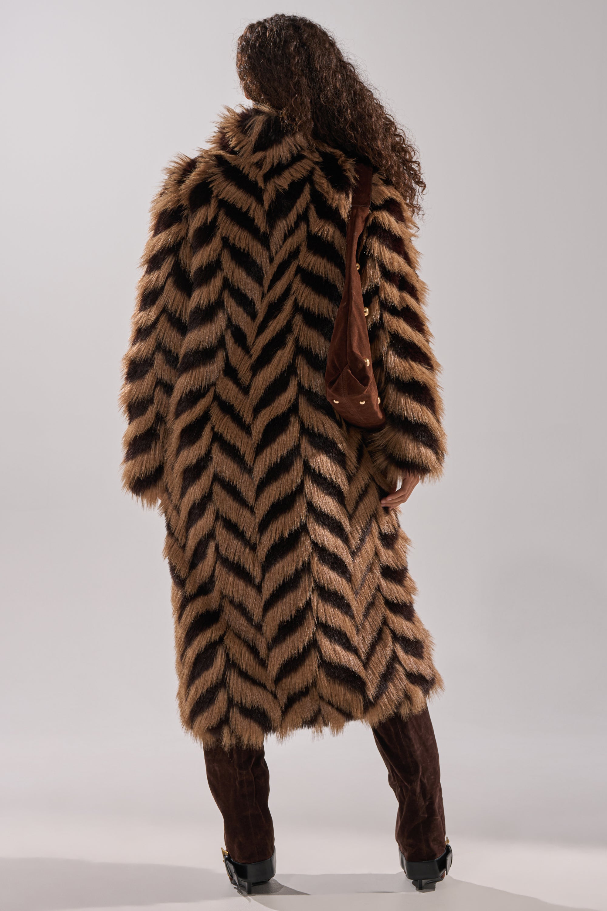 A person with long curly hair faces away, wearing the AUTUMN FAUX FUR COAT featuring a bold brown and black chevron pattern, paired with dark brown pants, black shoes, and a brown shoulder bag.