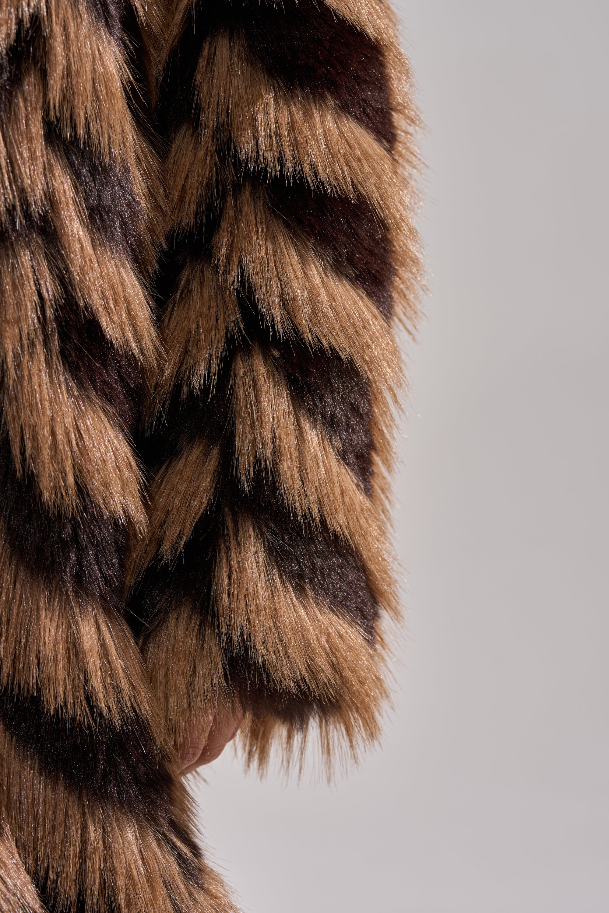 Close-up of a person wearing the AUTUMN FAUX FUR COAT in brown and black stripes, with their hand partially visible at the side, set against a plain gray background.