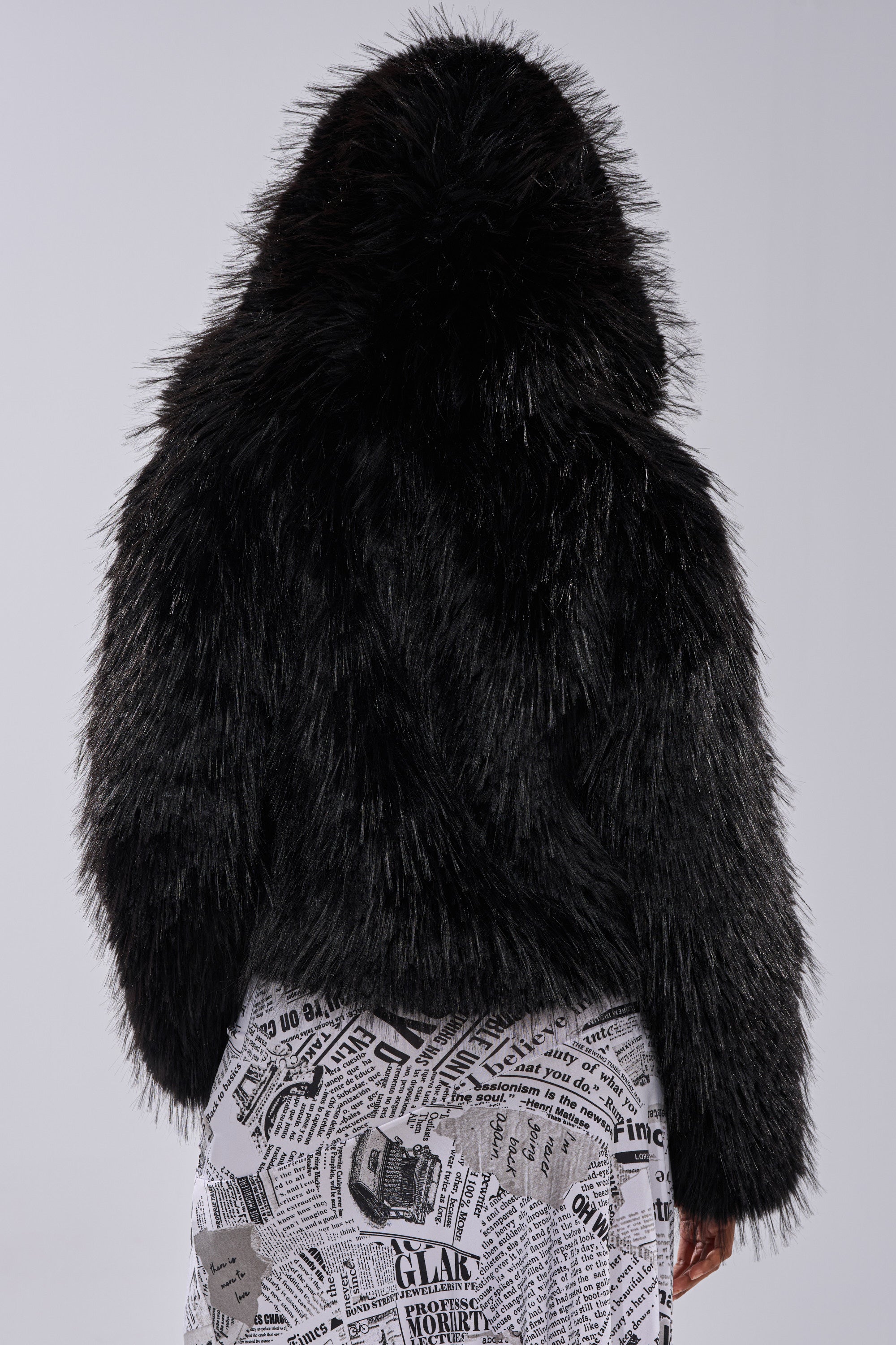 A person wearing the LOSING IT HOODED FAUX FUR jacket stands with their back to the camera, paired with white bottoms featuring a black newspaper print. The background is plain light gray.