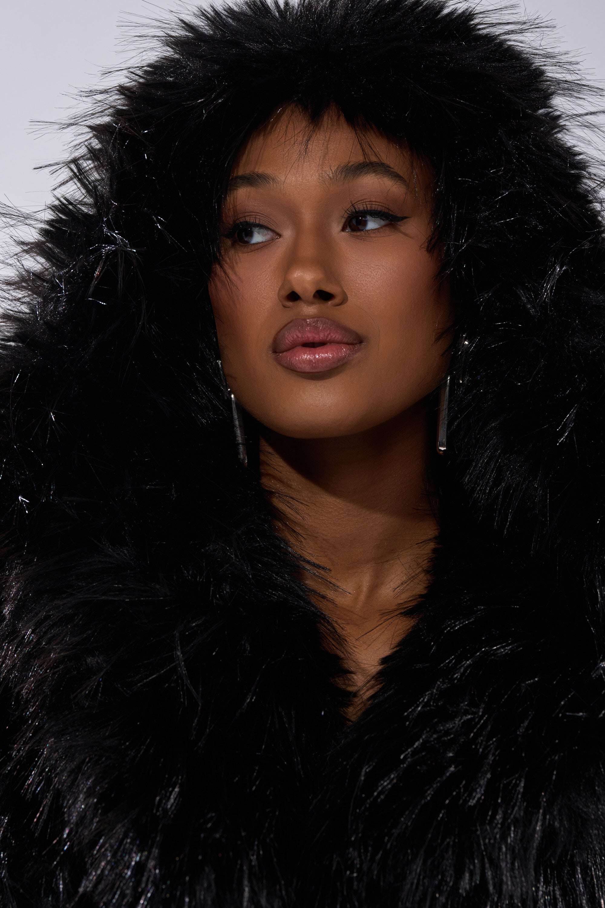 Model wears the LOSING IT HOODED FAUX FUR in black, gazing slightly to the side with a neutral expression and natural makeup against a plain light background.