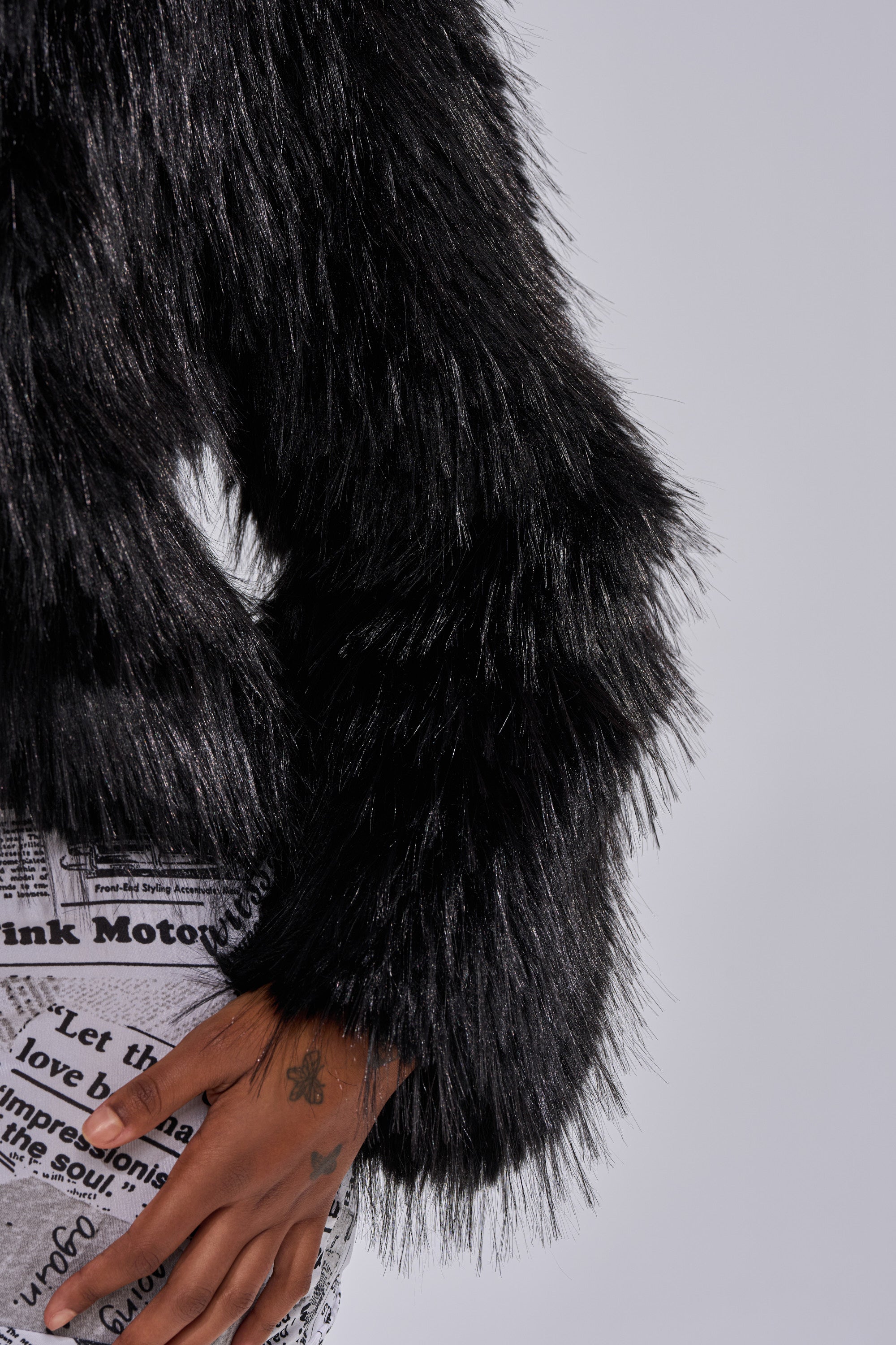 A person models the LOSING IT HOODED FAUX FUR jacket with newspaper print pants. Their tattooed hand rests at their side, set against a plain light background.