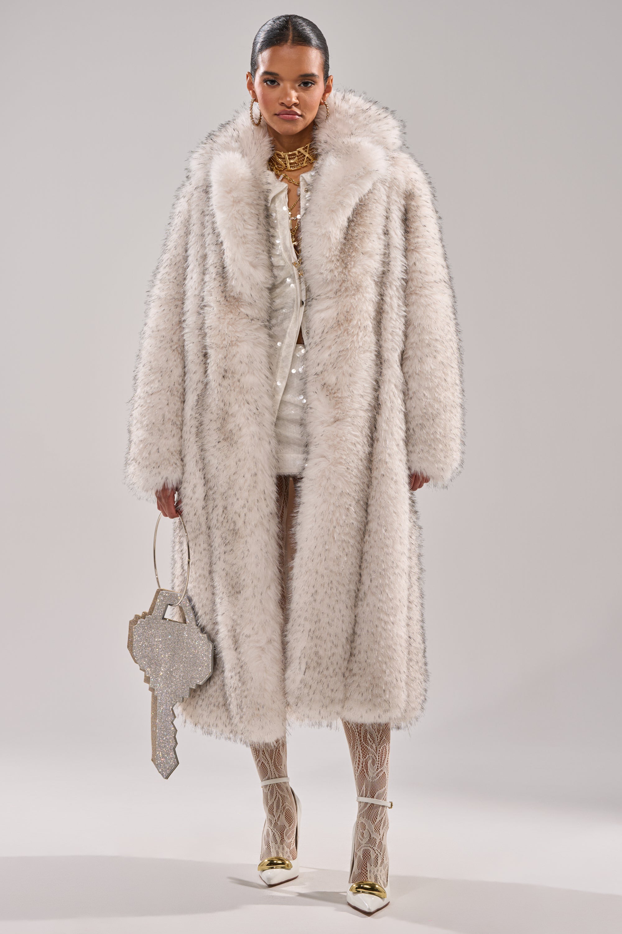 A person stands facing forward, wearing the HEAVY ON THE APRES FAUX FUR COAT over a white outfit. They hold a rhinestone key-shaped handbag and wear gold heels with white patterned tights.