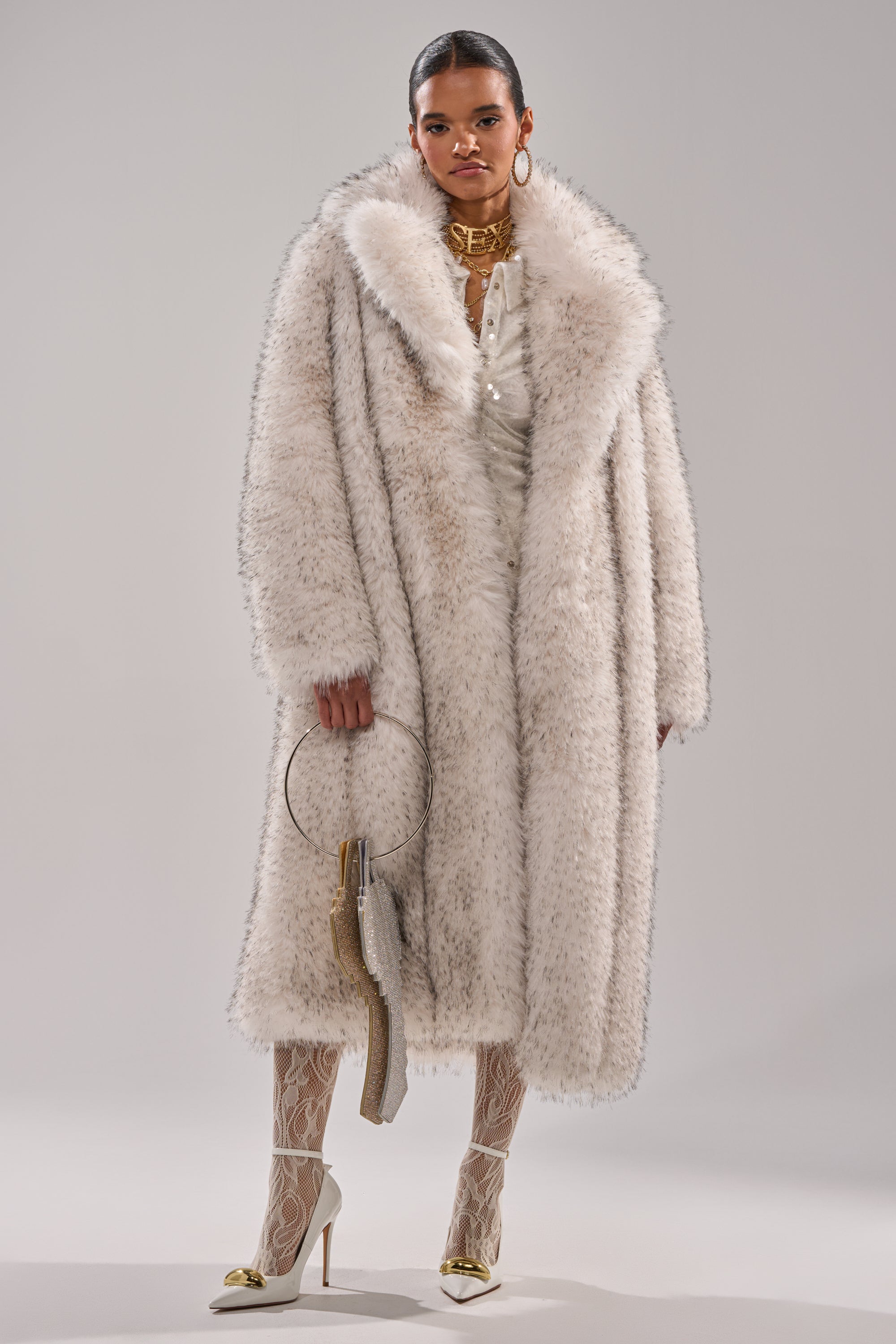 A woman exudes confidence in the HEAVY ON THE APRES FAUX FUR COAT, styled with white lace tights, heels, a circular-handle handbag, and bold gold jewelry—including a statement necklace—set against a plain backdrop.