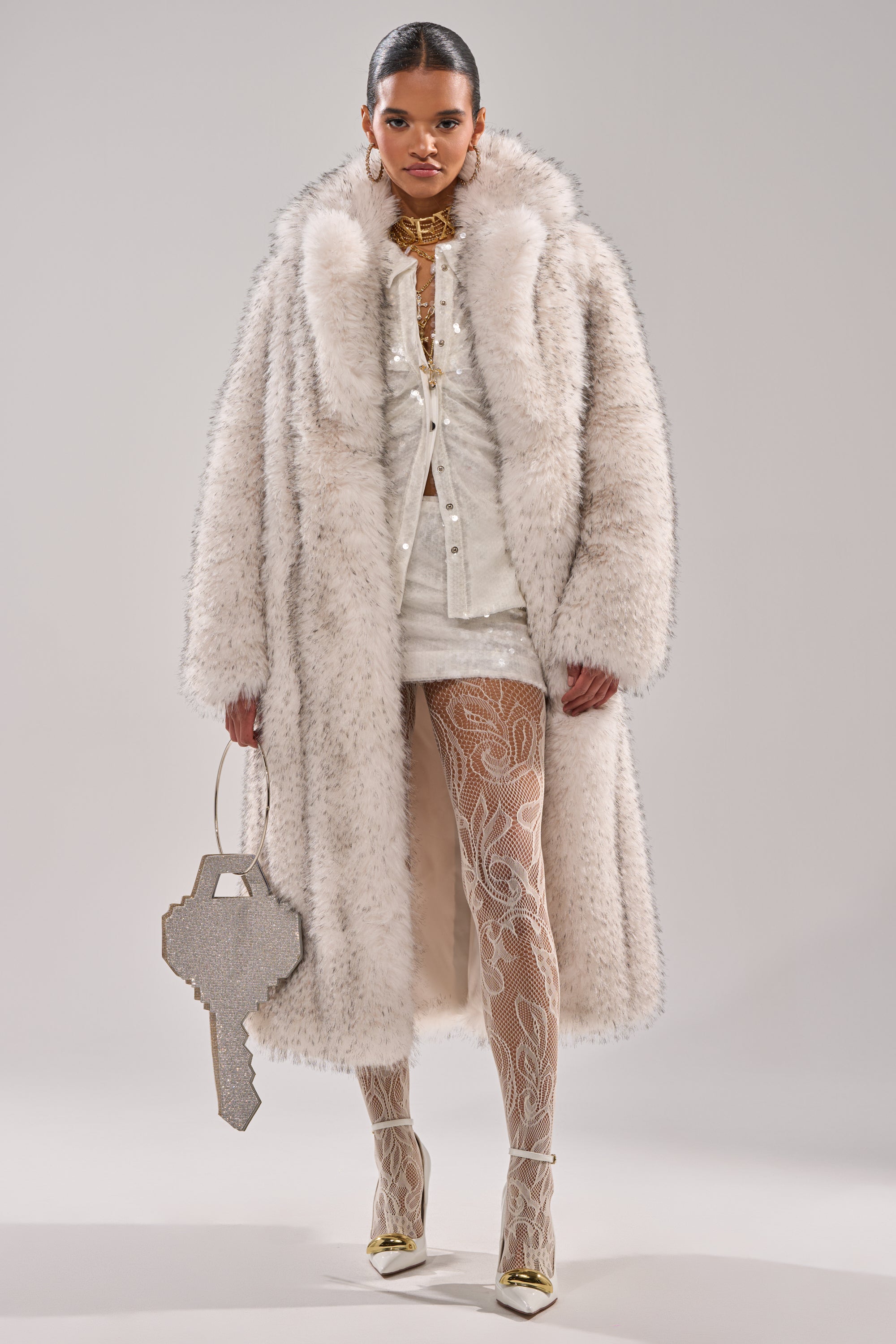 A model wears the HEAVY ON THE APRES FAUX FUR COAT over a white mini dress, with patterned tights and white heels with gold tips. She carries a key-shaped handbag, accessorized with bold gold jewelry, against a plain background.