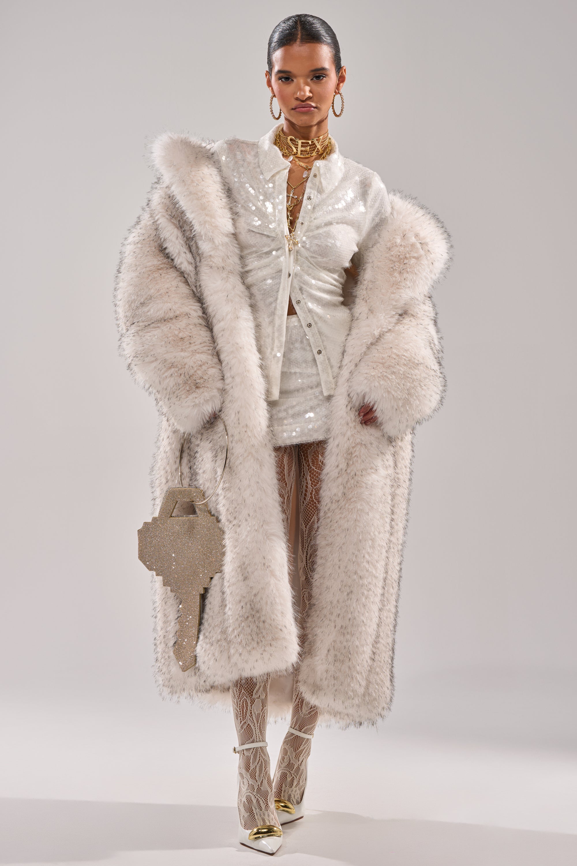 A woman models the HEAVY ON THE APRES FAUX FUR COAT over a sequined white mini dress, white lace tights, and gold pointed heels, accessorizing with a textured light-colored handbag and bold jewelry including a “SEX” choker.