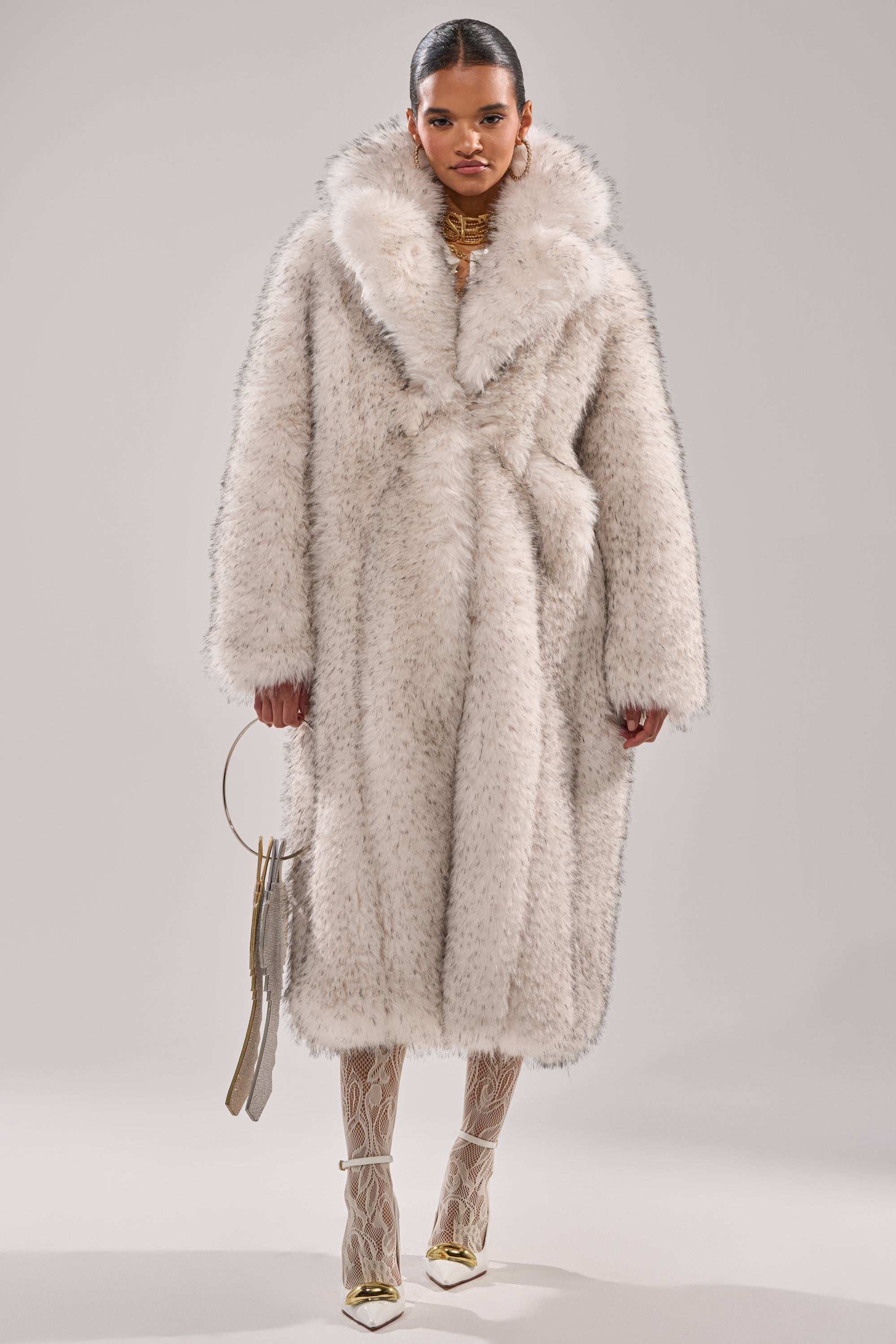 A woman stands confidently in the HEAVY ON THE APRES FAUX FUR COAT, its oversized white faux fur and large collar adding drama. She accessorizes with a small handbag, patterned tights, and gold pointed heels against a light gray background.