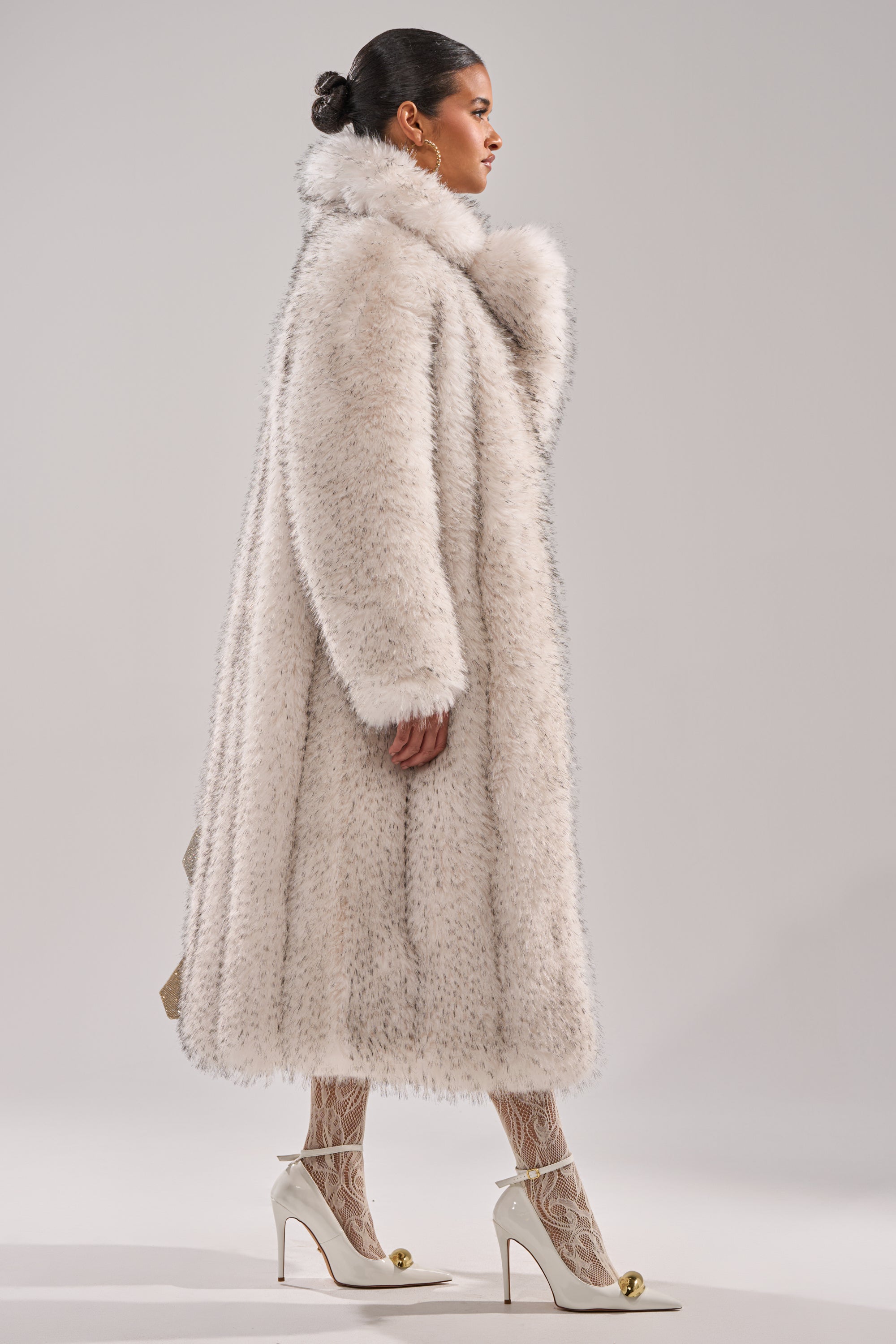 A woman with her hair in a bun wears the oversized white HEAVY ON THE APRES FAUX FUR COAT, paired with white patterned tights and white high heels with gold tips, standing in profile against a plain light background.