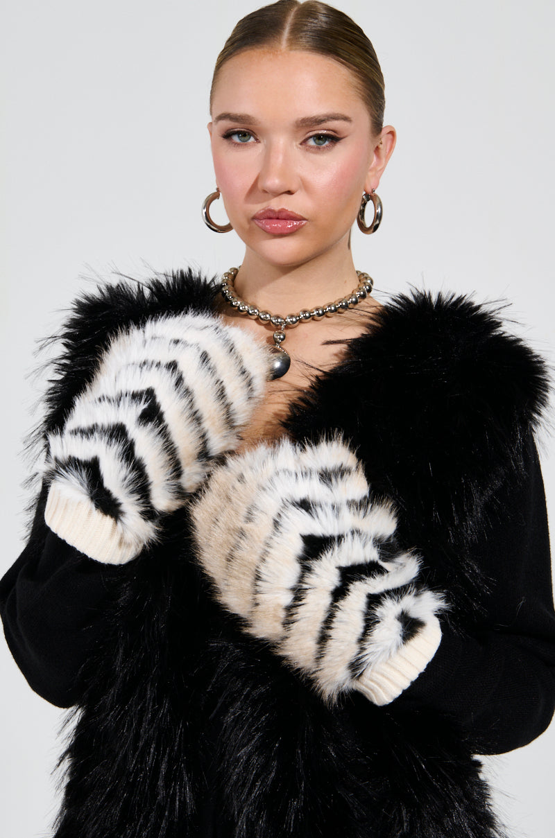A woman wearing a furry black vest and large hoop earrings poses confidently with her hands up, showcasing her WILDER FAUX FUR MITTENS featuring a chevron striped pattern. She has a neutral expression and her hair is pulled back.