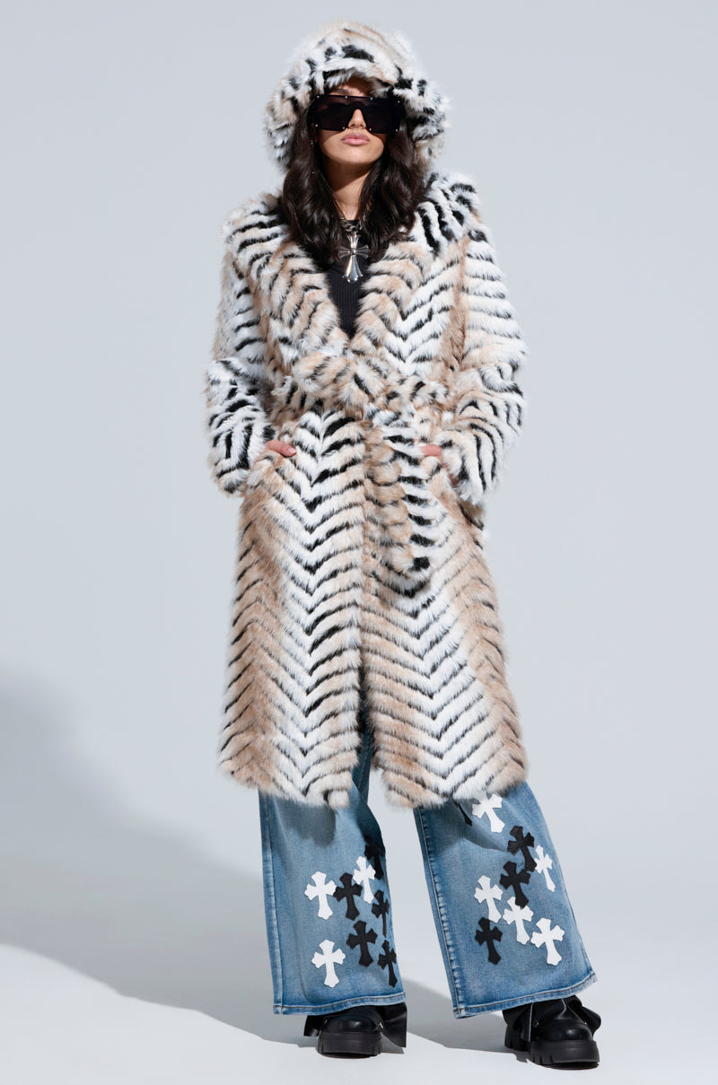 Dressed in the LOLA HOODED FAUX FUR JACKET with its oversized tiger-striped pattern, the individual pairs it with large sunglasses and wide-leg jeans adorned with cross designs, standing against a plain background.