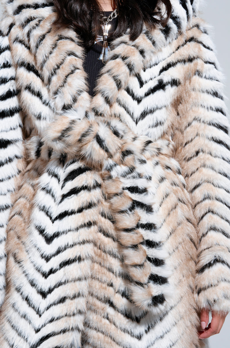A person is dressed in the LOLA HOODED FAUX FUR JACKET, showcasing its striking black, white, and beige zigzag pattern. This thick and luxuriously soft coat features an oversized longline silhouette with a tie at the waist for a stylish and cozy appearance. The plain background accentuates the jacket's bold design.