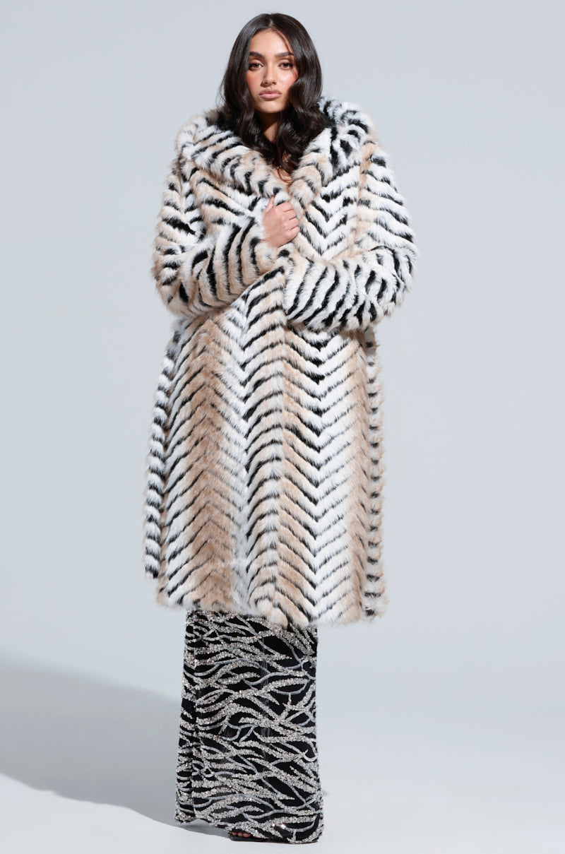 A person wearing a LOLA HOODED FAUX FUR JACKET, featuring an oversized longline design with tiger stripes, over a long dress adorned with a wavy pattern poses against a plain background.
