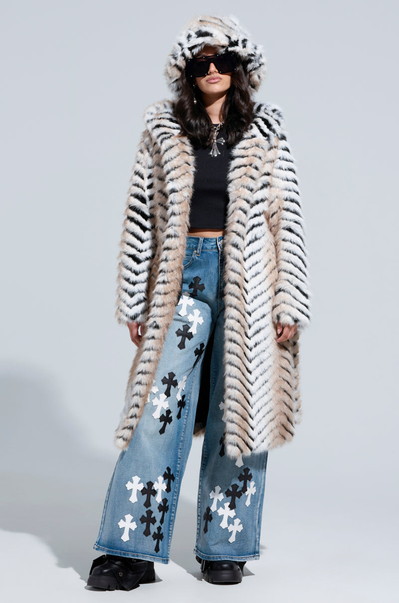 A person exudes confidence in an oversized LOLA HOODED FAUX FUR JACKET with a tiger-striped pattern and a matching hat. Large black sunglasses, paired with a black top and wide-legged jeans featuring black and white cross patterns, complete their look against a simple gray background.