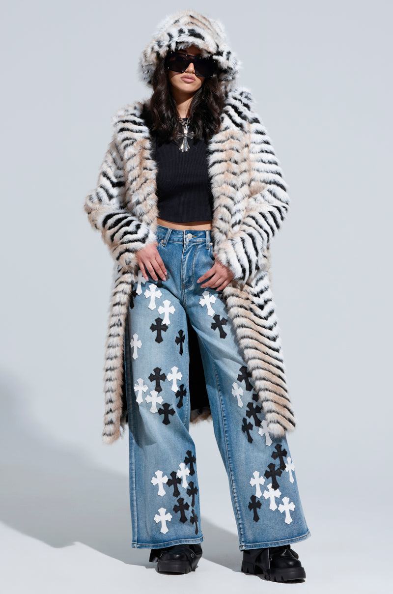 Sporting the LOLA HOODED FAUX FUR JACKET in a bold tiger-striped design, a person stylishly layers it over a black sweater. Complementing their look with wide-legged jeans adorned with white and black cross patterns and finishing off with black platform shoes, they pose confidently against a plain backdrop, radiating easy elegance.