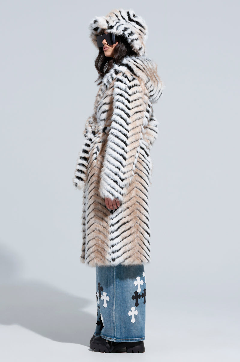 A person is wrapped in a LOLA HOODED FAUX FUR JACKET with tiger stripes, paired with a matching hat, sunglasses, and blue jeans featuring white cross patterns as they stand against a plain background.
