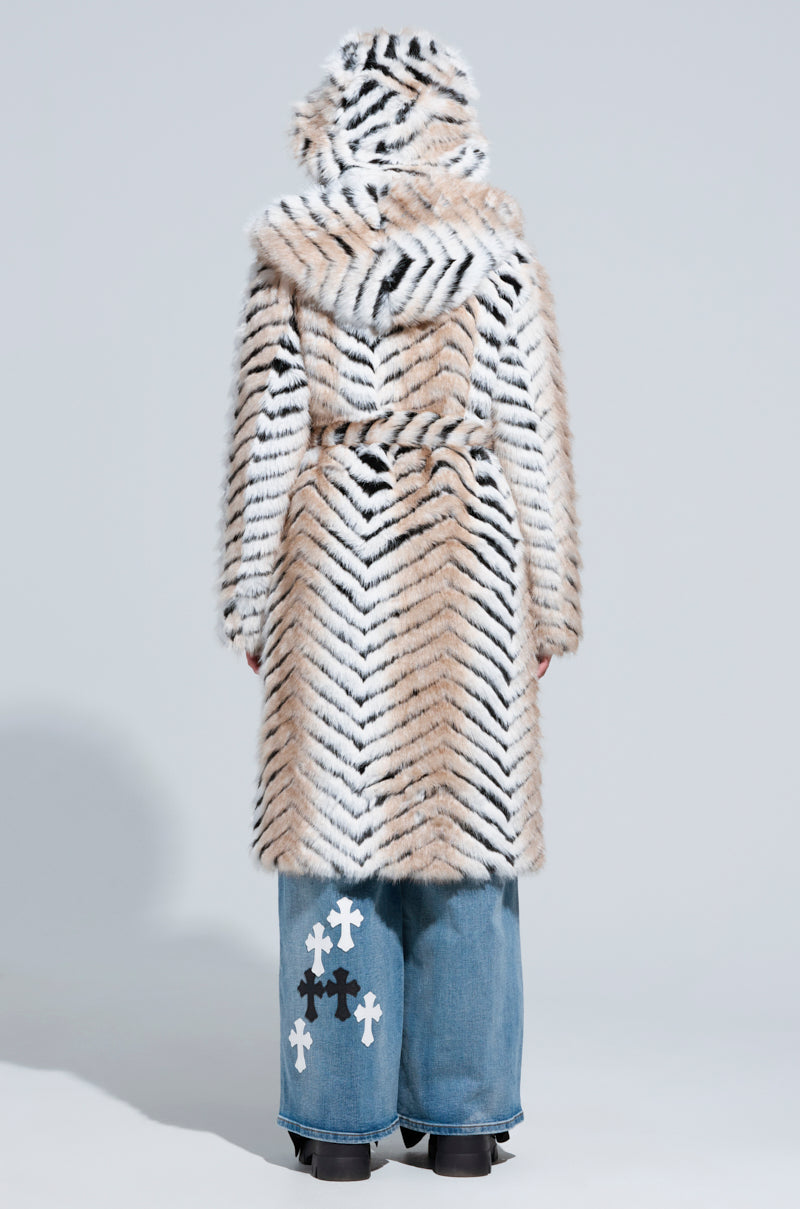 A person stands with their back to the camera, draped in an oversized longline coat featuring a tiger-striped pattern. They're wearing wide-leg jeans adorned with white cross designs on one leg and black shoes, while the hood of their LOLA HOODED FAUX FUR JACKET is pulled up. The background is plain gray.