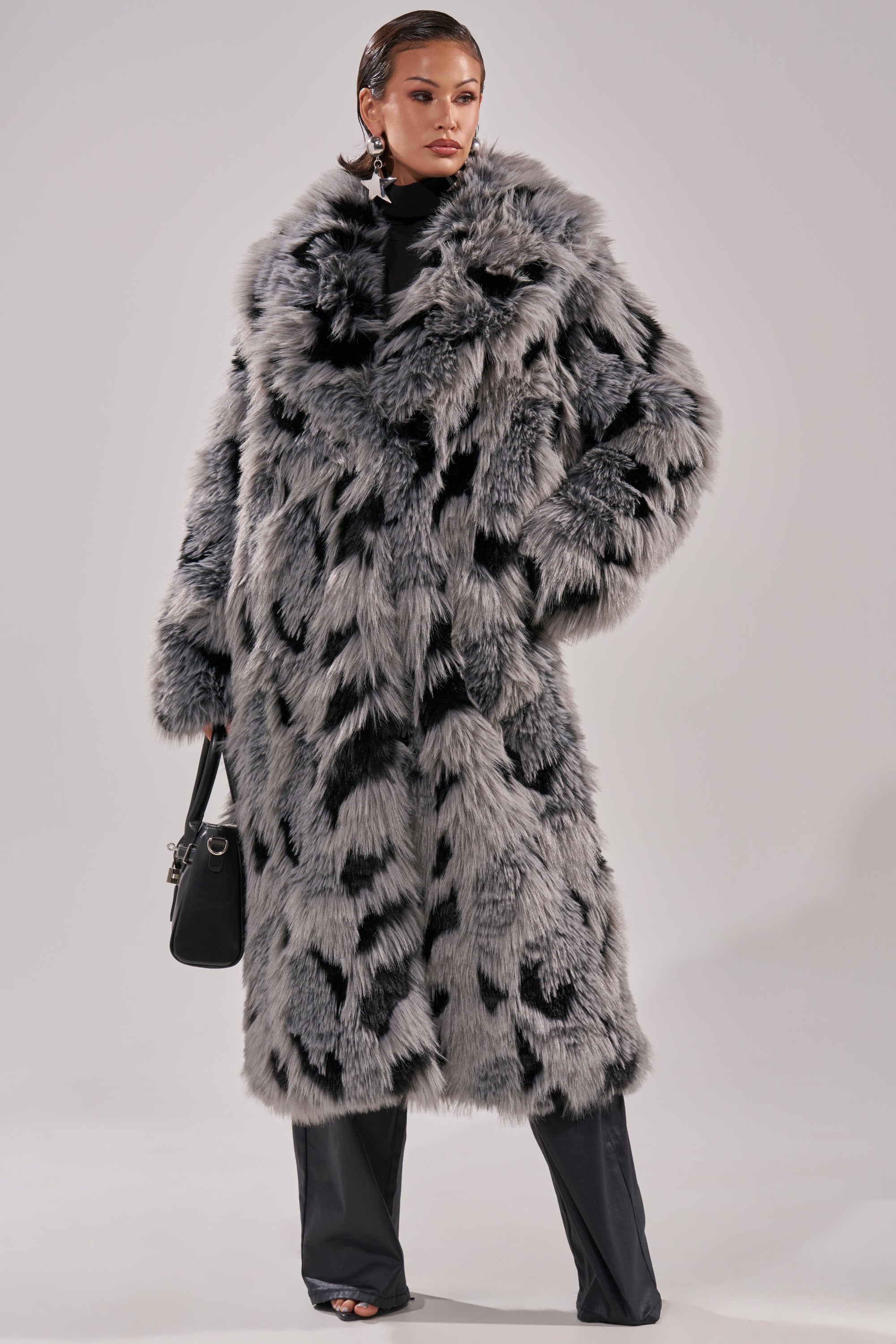 A woman models the APRES SKI FAUX FUR COAT, featuring a longline silhouette and black and gray spots. She pairs it with black pants, a black top, statement earrings, and carries a small black handbag.