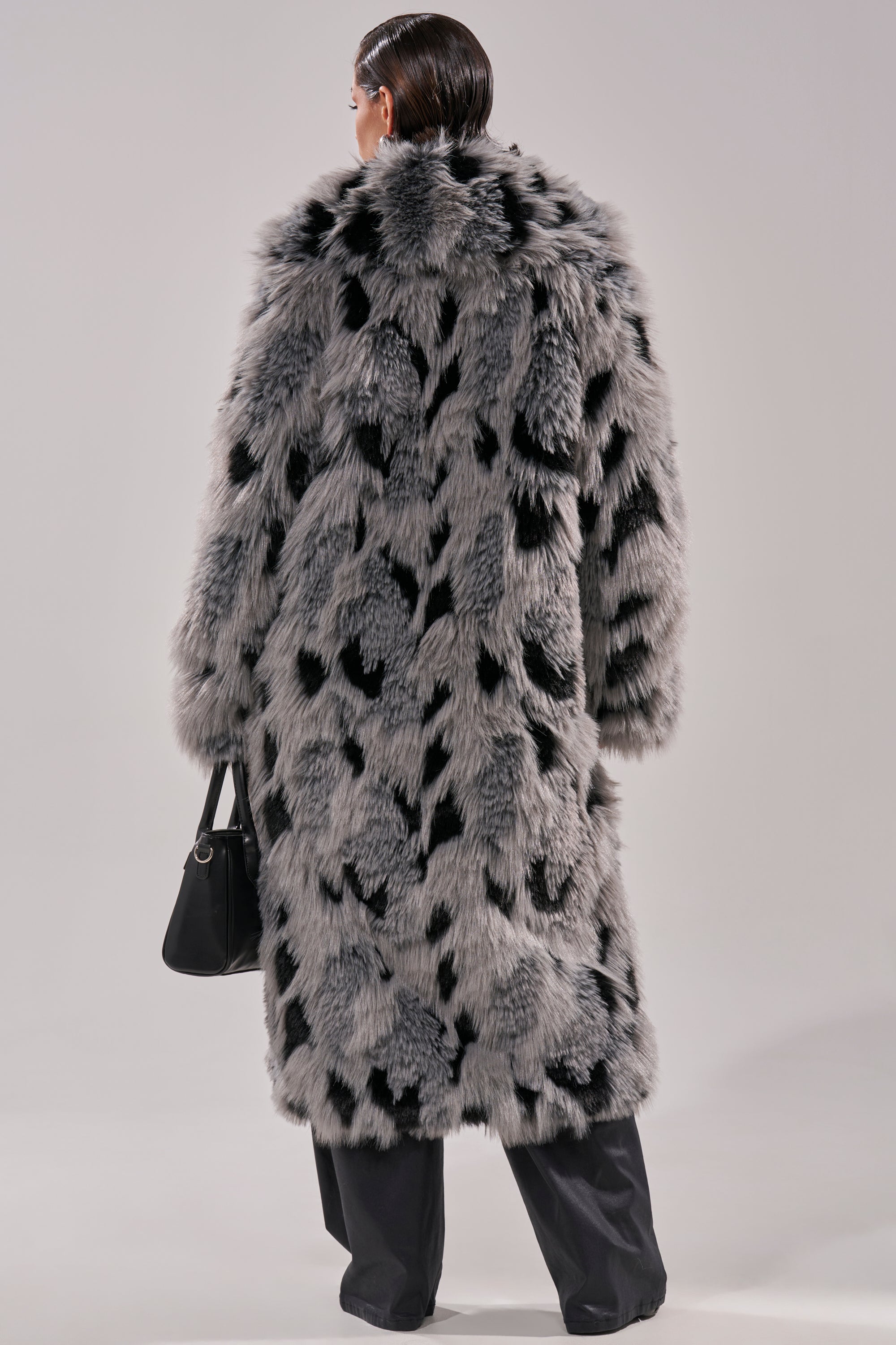 A person with slicked-back hair showcases the APRES SKI FAUX FUR COAT in black and gray spots, featuring a longline silhouette. Paired with dark trousers and a small black handbag, they’re seen from the back against a plain background.