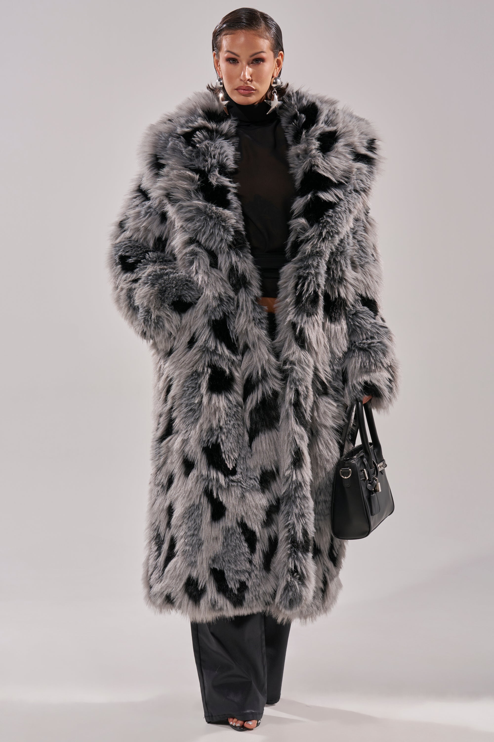 A person wearing the APRES SKI FAUX FUR COAT, featuring a longline silhouette with black and gray spots, over black clothing and holding a black handbag, stands against a plain light background.