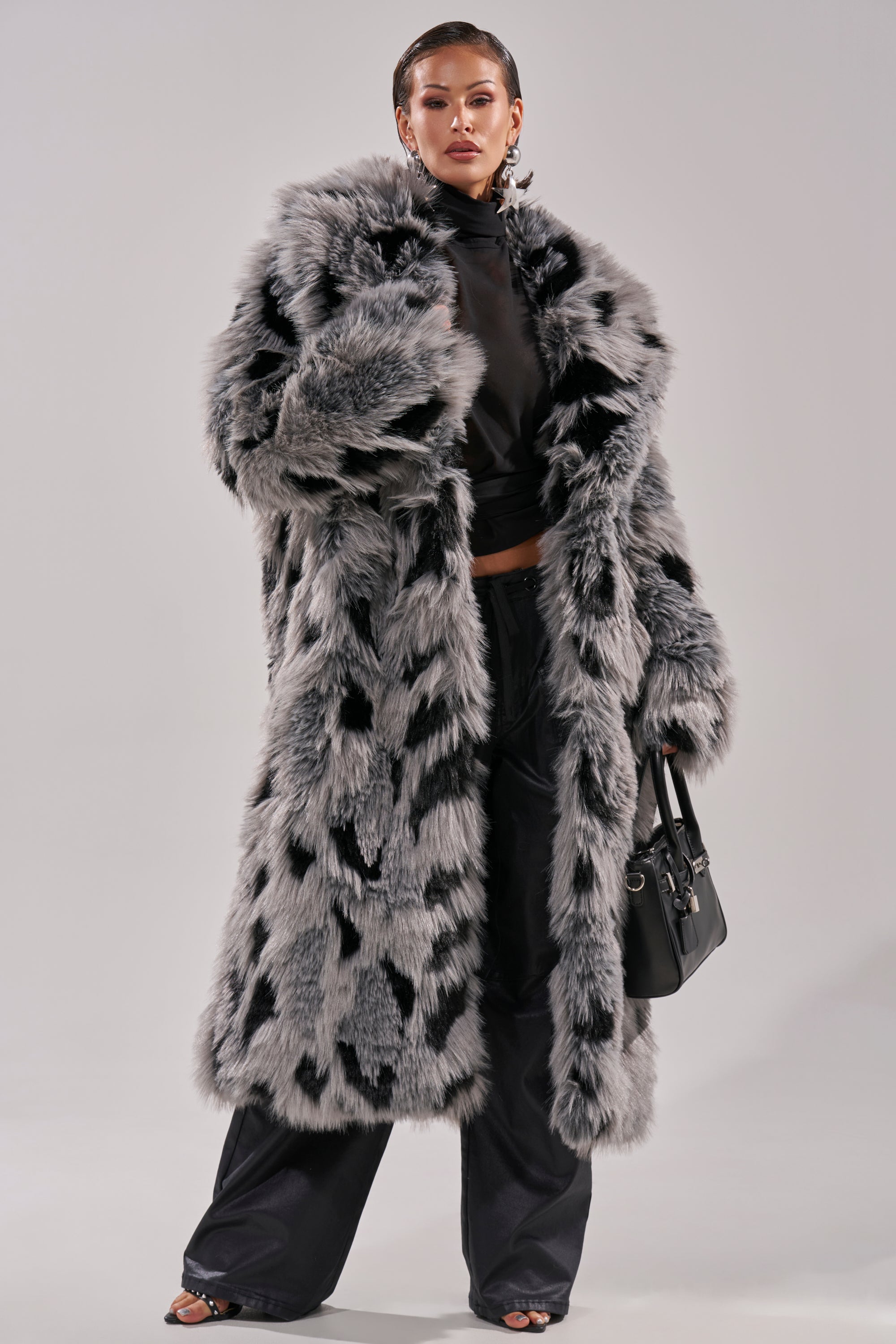 A person stands confidently in the APRES SKI FAUX FUR COAT, featuring a longline gray and black spotted faux fur design over black pants and a turtleneck, holding a black handbag against a plain light background.