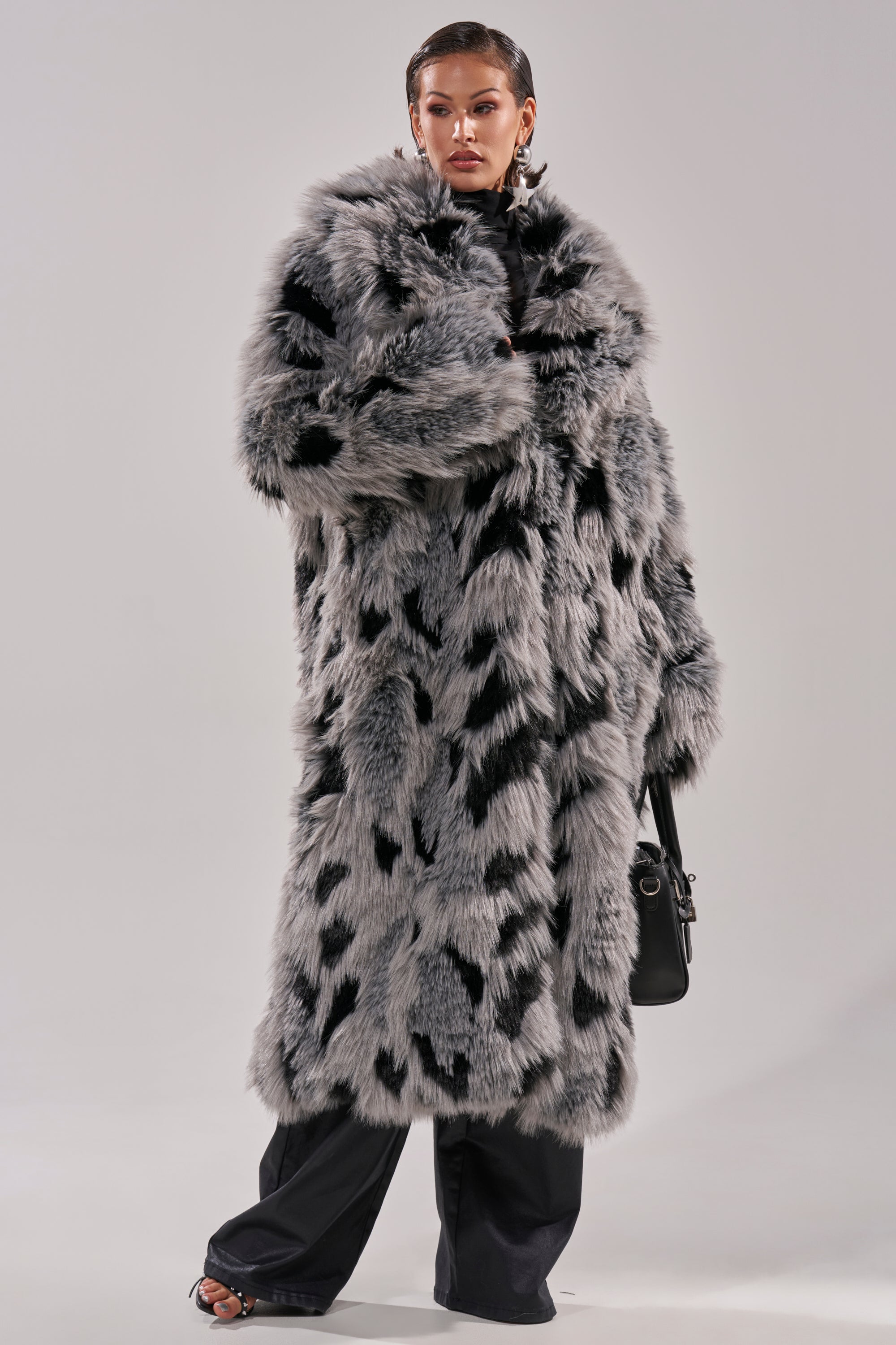 A woman models the APRES SKI FAUX FUR COAT—a gray longline faux fur coat with black spots—styled with black pants, open-toe heels, statement earrings, and a black handbag against a plain light background.