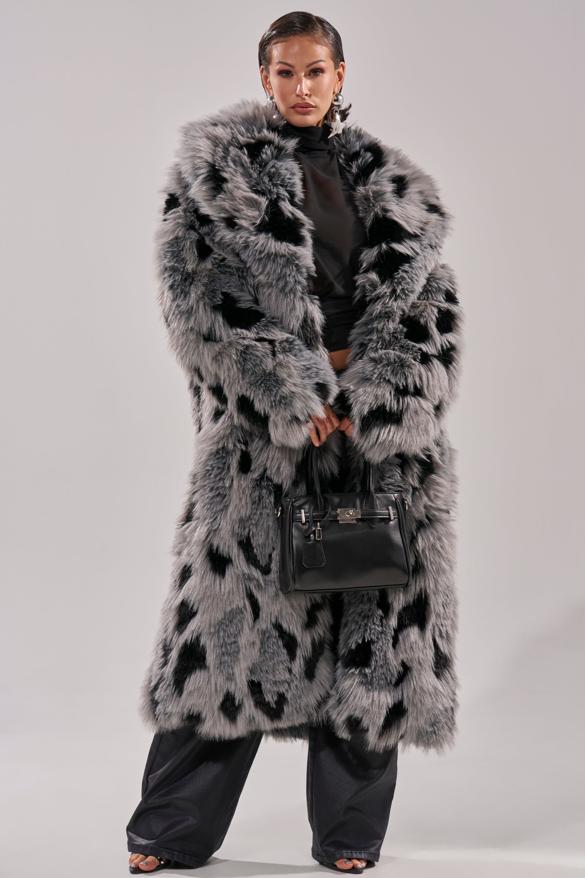 A person confidently stands against a plain light background wearing the APRES SKI FAUX FUR COAT in a longline black and gray spotted design, paired with black pants and holding a black handbag.