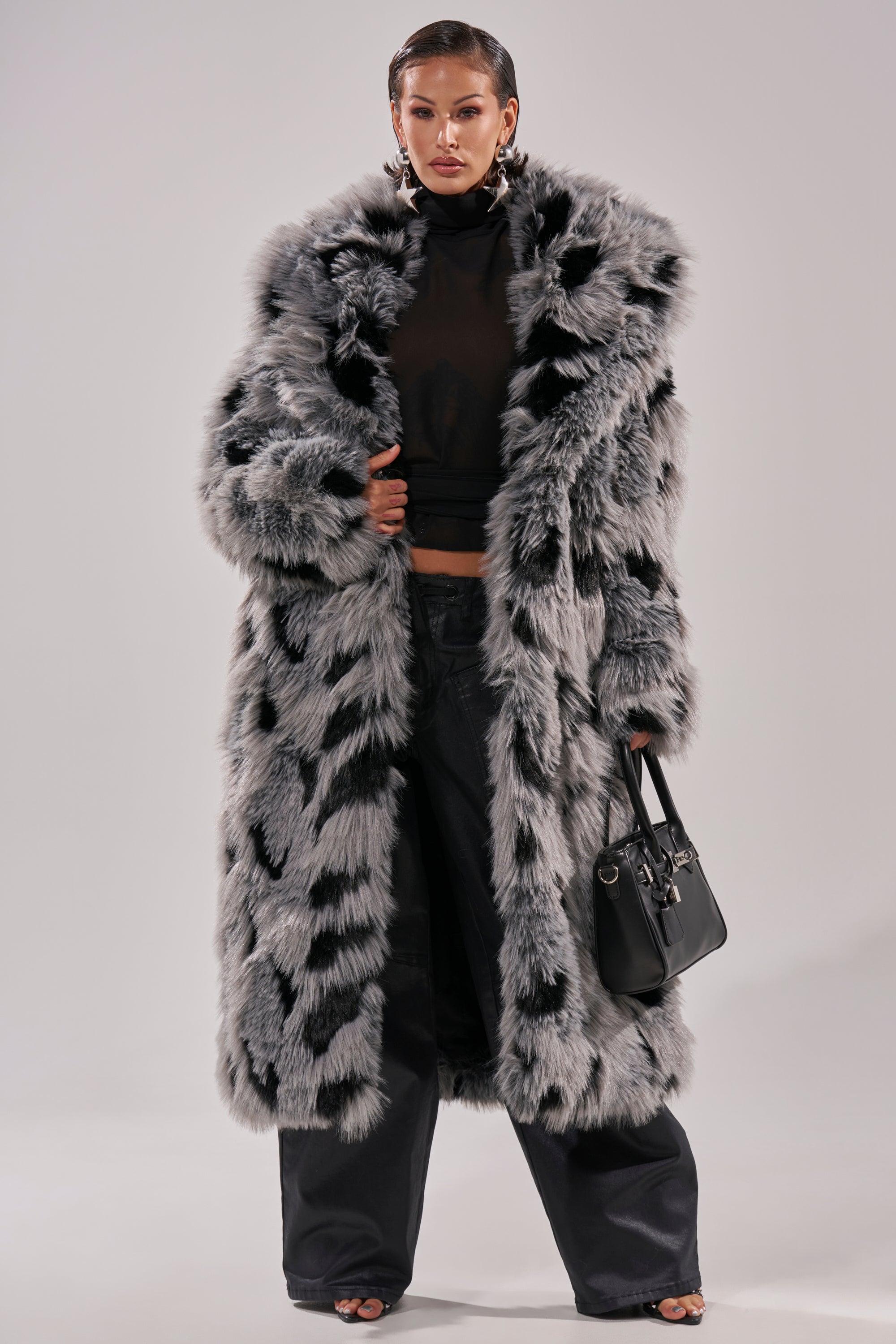 Wearing the APRES SKI FAUX FUR COAT, a person pairs it with a black sheer turtleneck, wide-leg black pants, and a black handbag, standing confidently against a plain background.