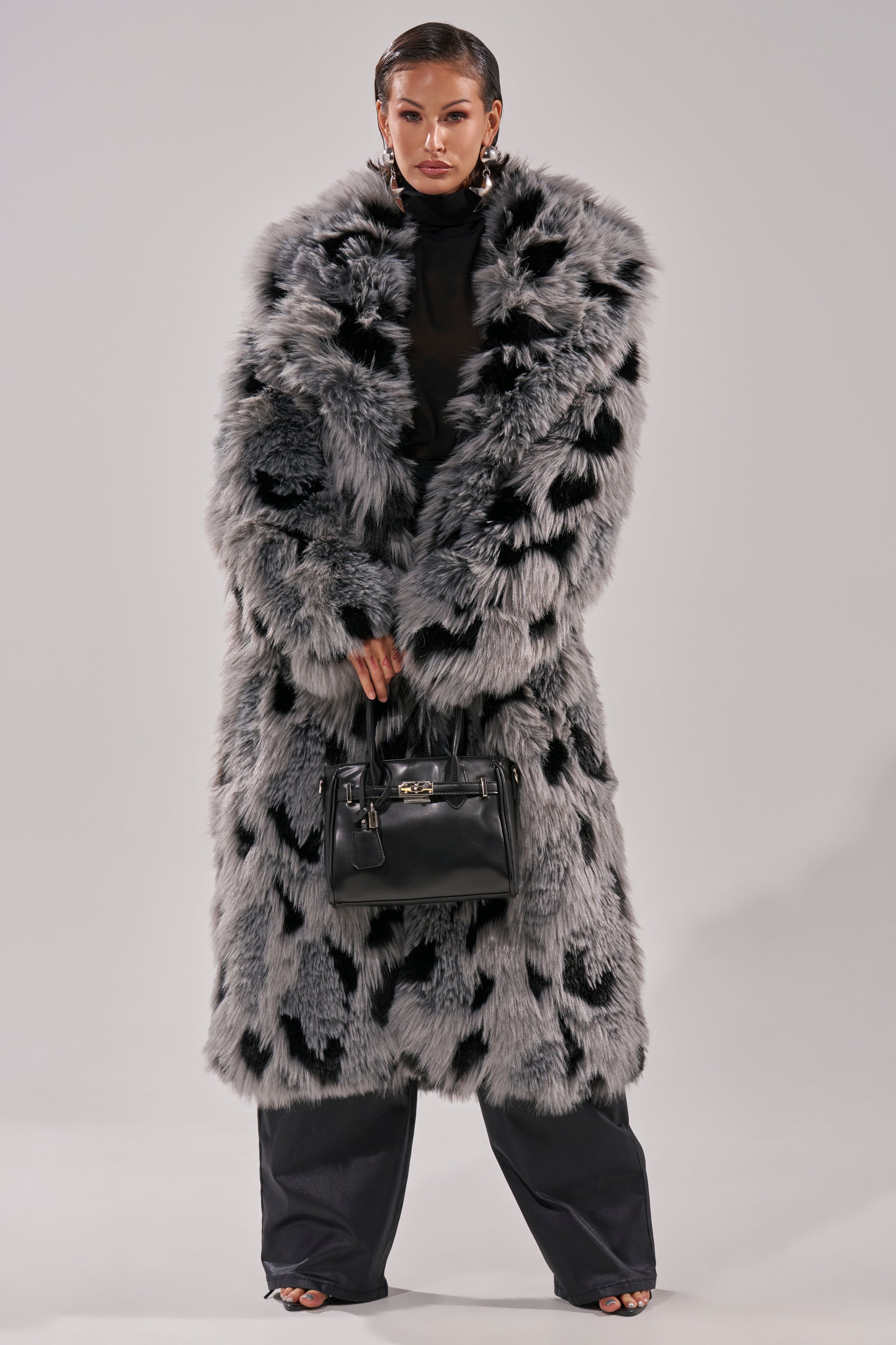 A person models the APRES SKI FAUX FUR COAT, featuring a long, oversized silhouette with black and gray spots. Styled with black pants, open-toe heels, statement earrings, a black handbag, and slicked-back hair for a dramatic look.