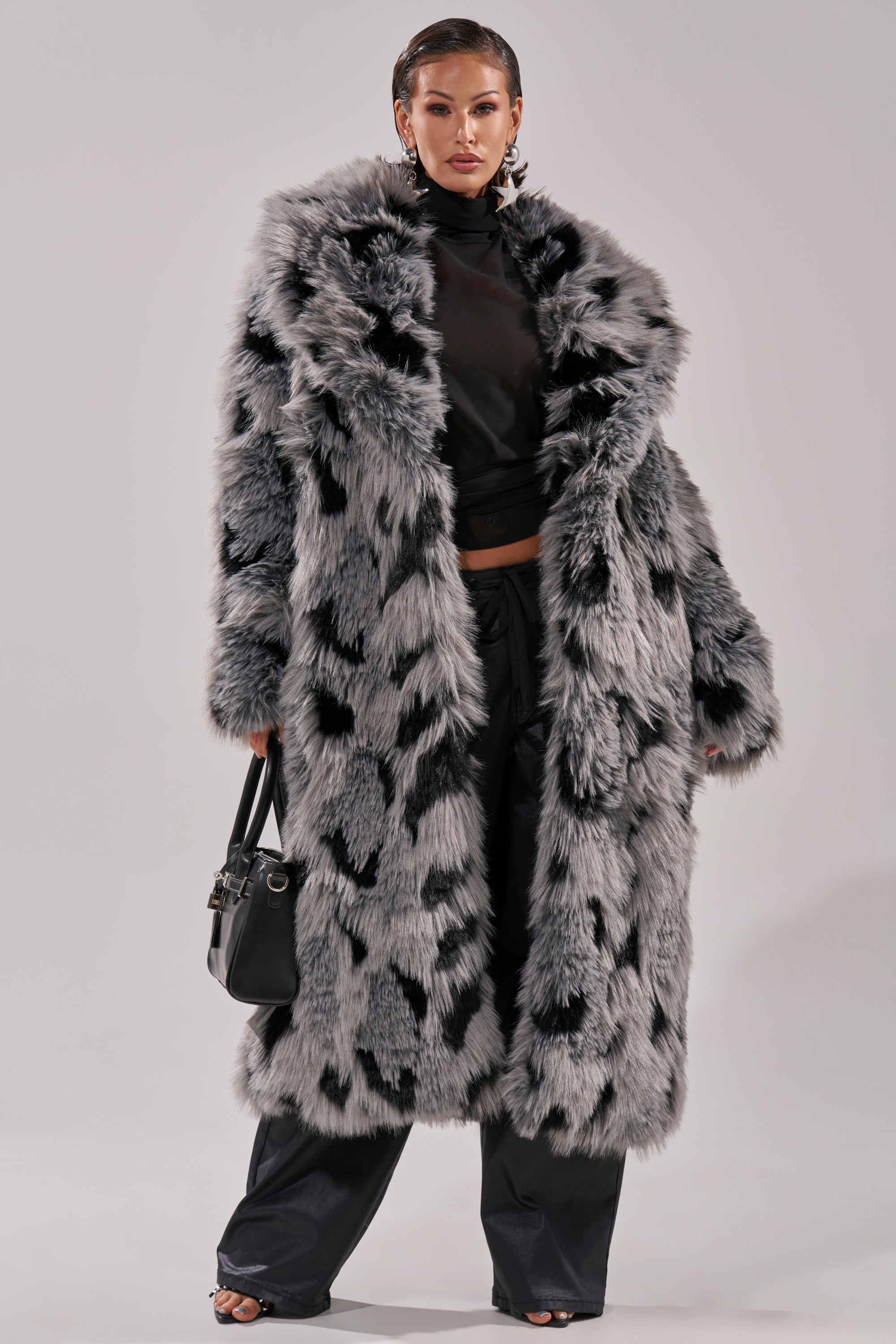 A woman exudes bold style in the APRES SKI FAUX FUR COAT, a longline silhouette with gray and black tones. With slicked-back hair, dark pants, black turtleneck, and a small handbag, she stands confidently against a plain background.