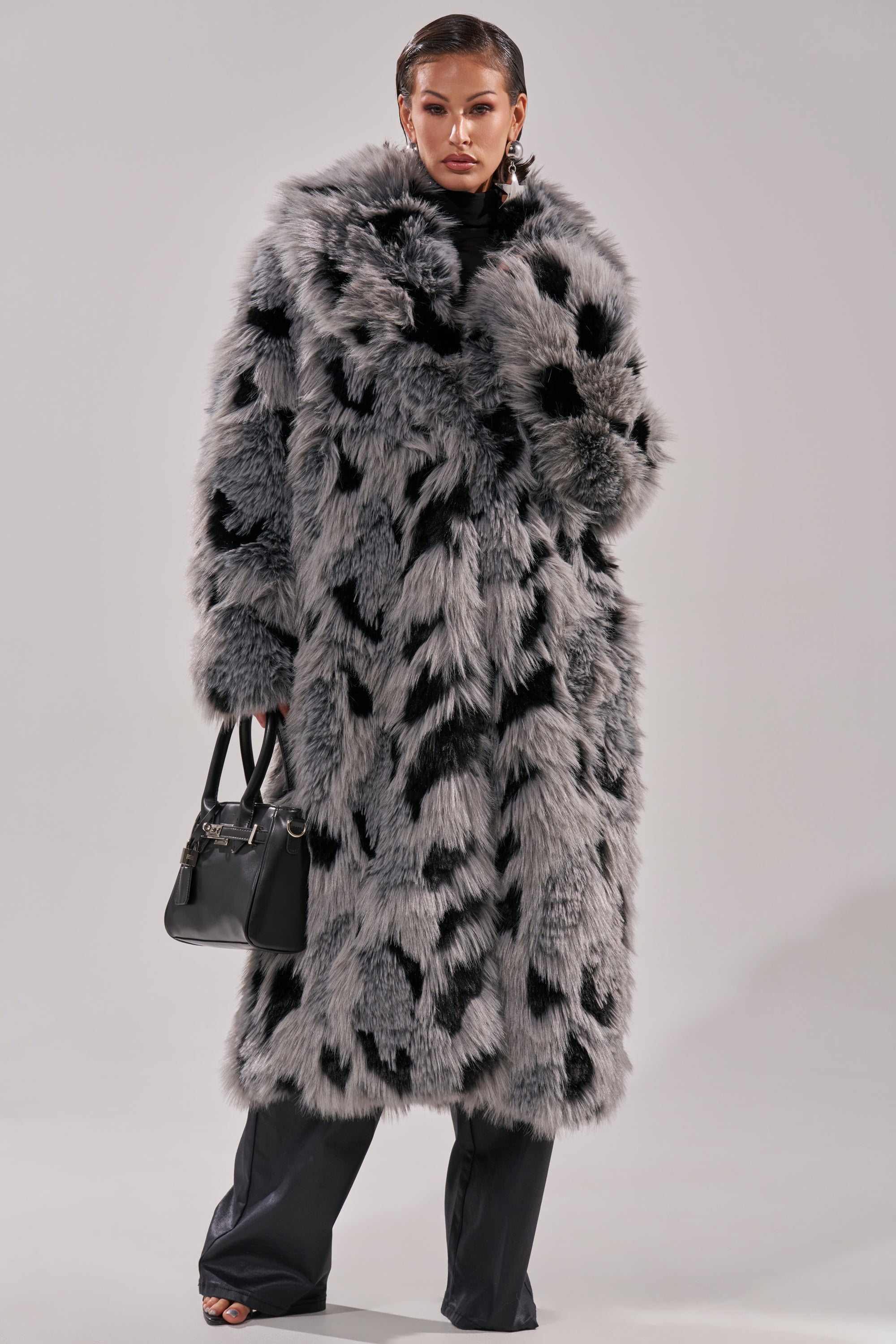 A person wears the APRES SKI FAUX FUR COAT, an oversized black-and-gray longline silhouette with black spots, paired with dark pants and a black handbag, standing against a plain light gray background.