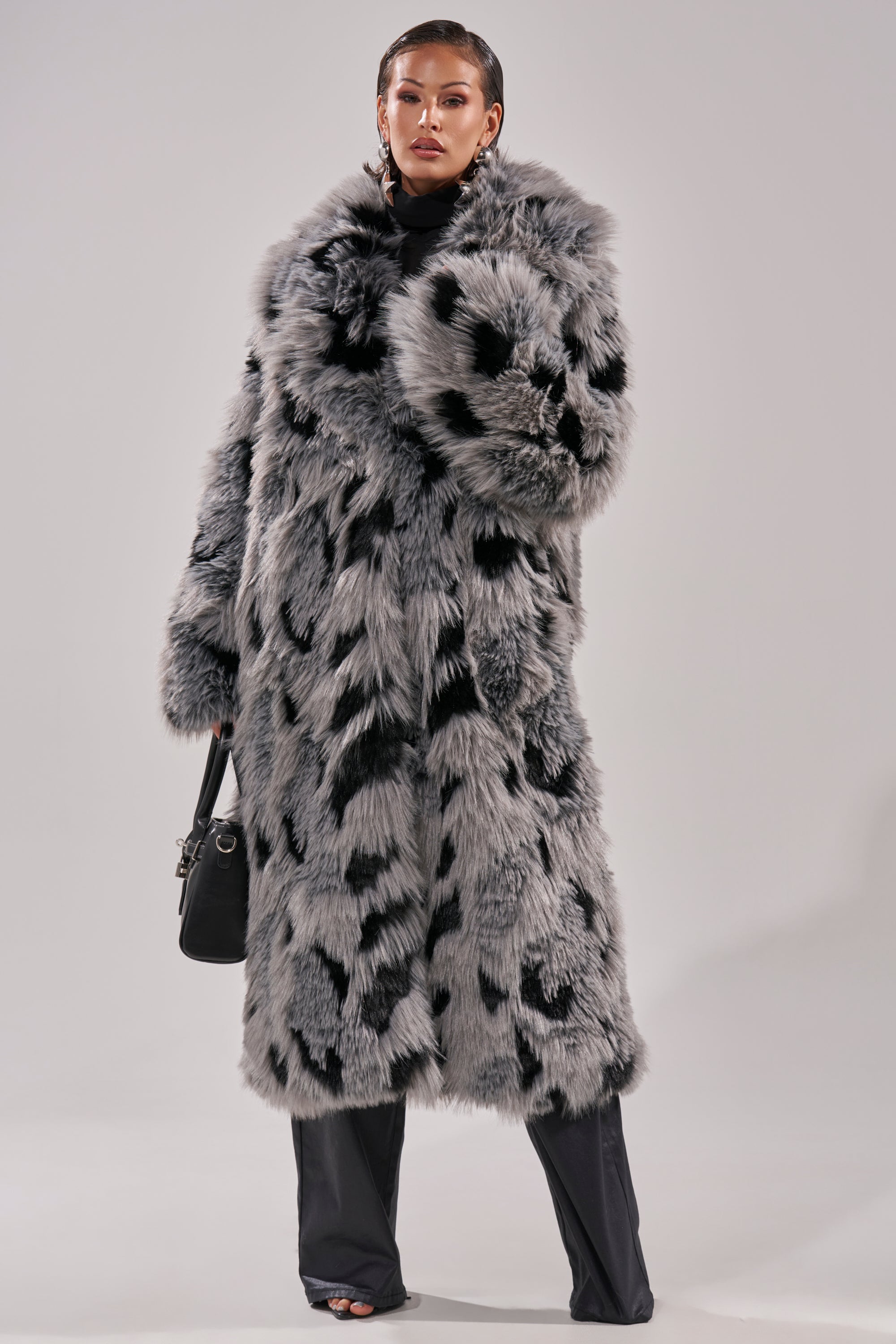 A person poses confidently in the APRES SKI FAUX FUR COAT, a longline gray faux fur coat with black spots. They style it with black pants, open-toe shoes, a small black handbag, and slicked-back hair against a plain light background.