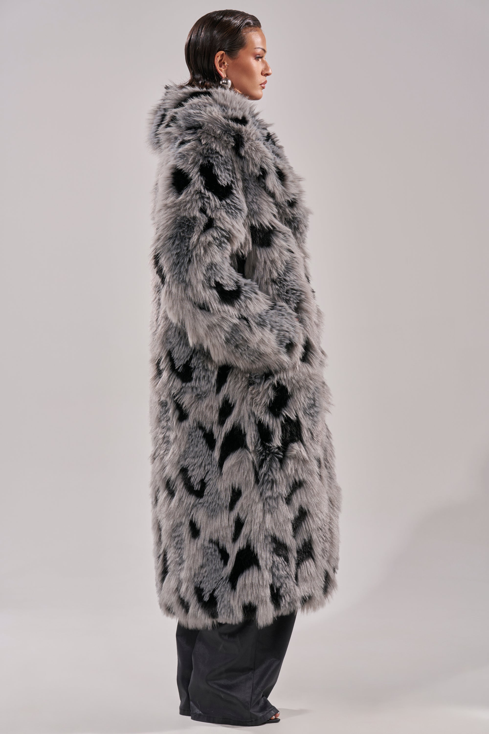 A person stands in profile against a light gray background, wearing the APRES SKI FAUX FUR COAT in a longline gray and black faux fur silhouette, paired with black pants and slicked-back hair.