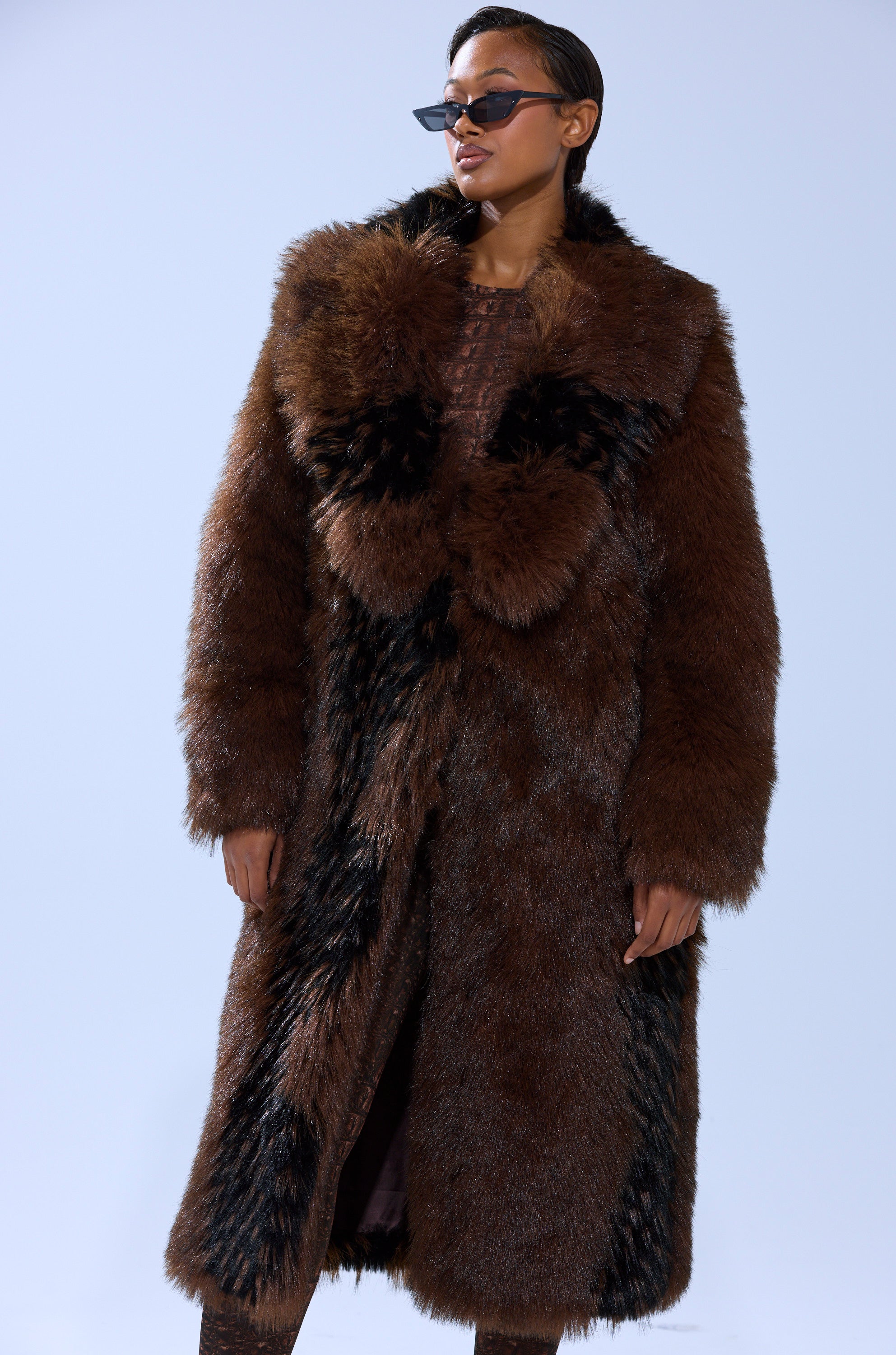 A person stands confidently against a plain light background, wearing sunglasses and the TELLURIDE FAUX FUR IN BROWN, a longline coat with dark accents.